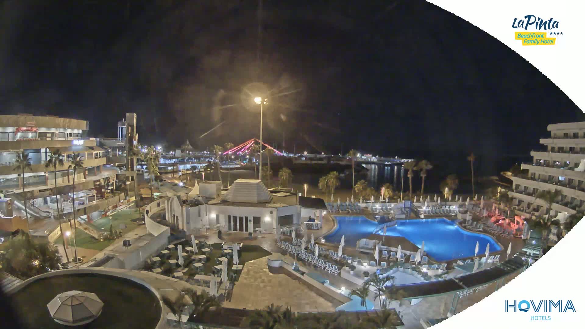 A nighttime aerial view shows a resort with swimming pools and surrounding buildings, overlooking a calm body of water and a bridge illuminated with lights.