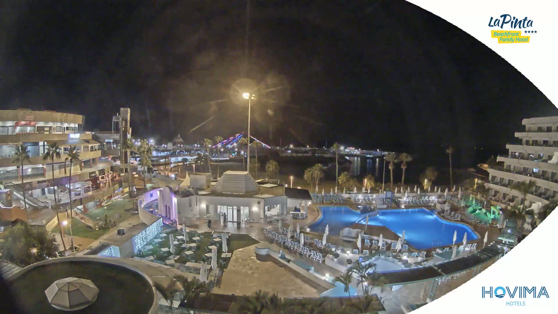 A nighttime aerial view of a resort complex with a swimming pool, illuminated buildings, and palm trees, overlooking a calm body of water and a brightly lit bridge in the distance.
