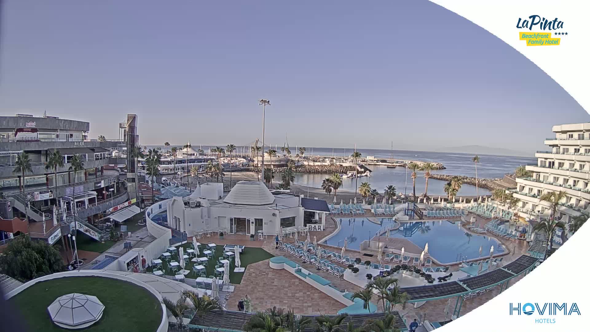 An elevated view shows a resort pool area, shops, and a marina on a clear day with a calm ocean in the background.