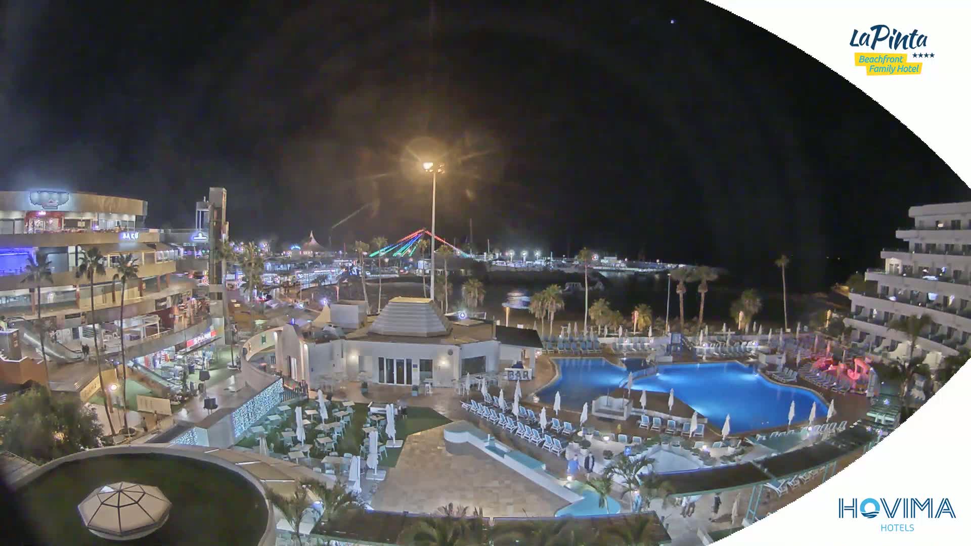 A nighttime aerial view of a resort area with illuminated buildings, palm trees, a swimming pool, and a brightly lit bridge in the distance under a clear, dark sky.