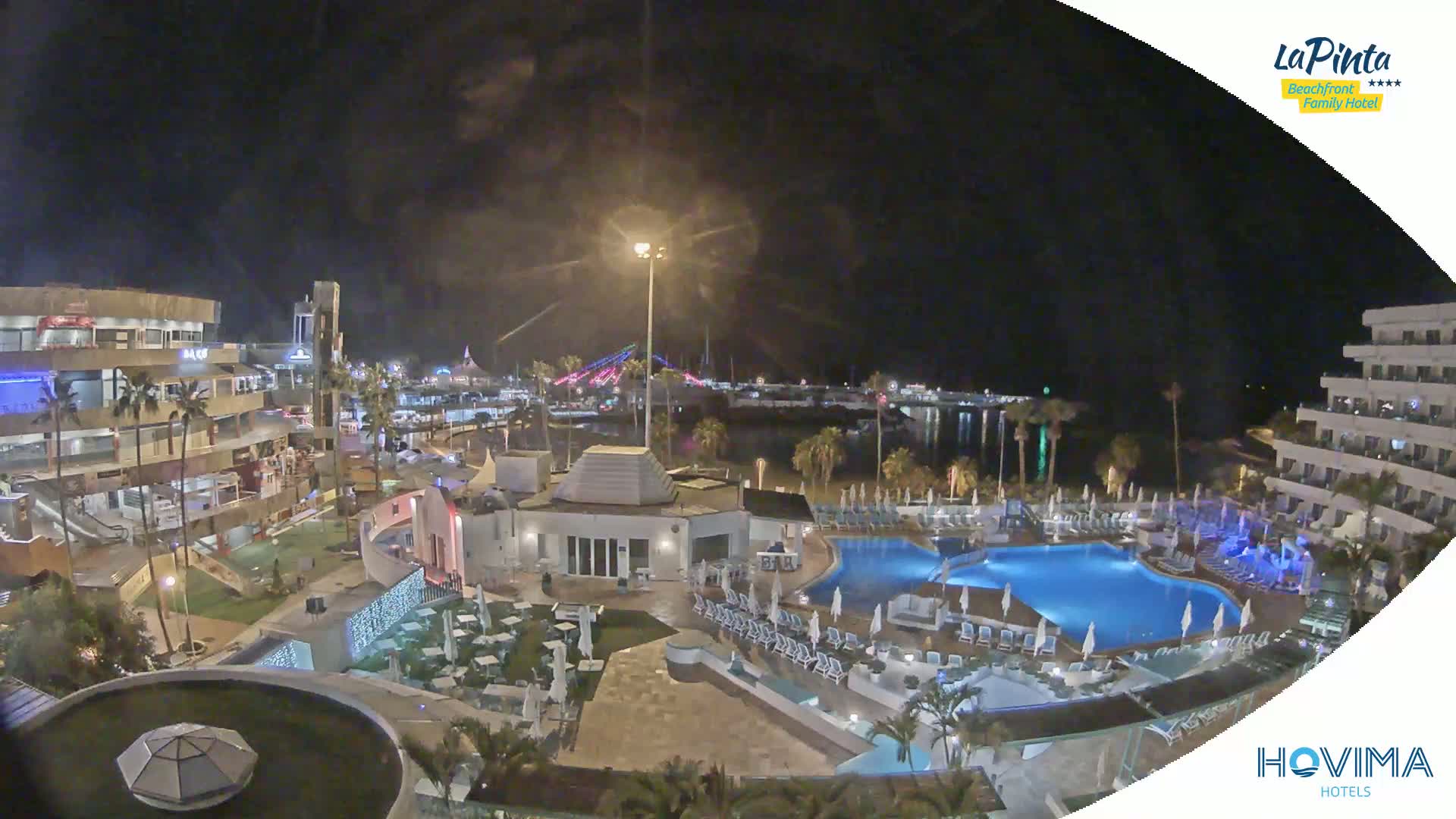 A nighttime view of a resort area with pools and buildings, illuminated by artificial lights, overlooking a calm body of water and a distant bridge.