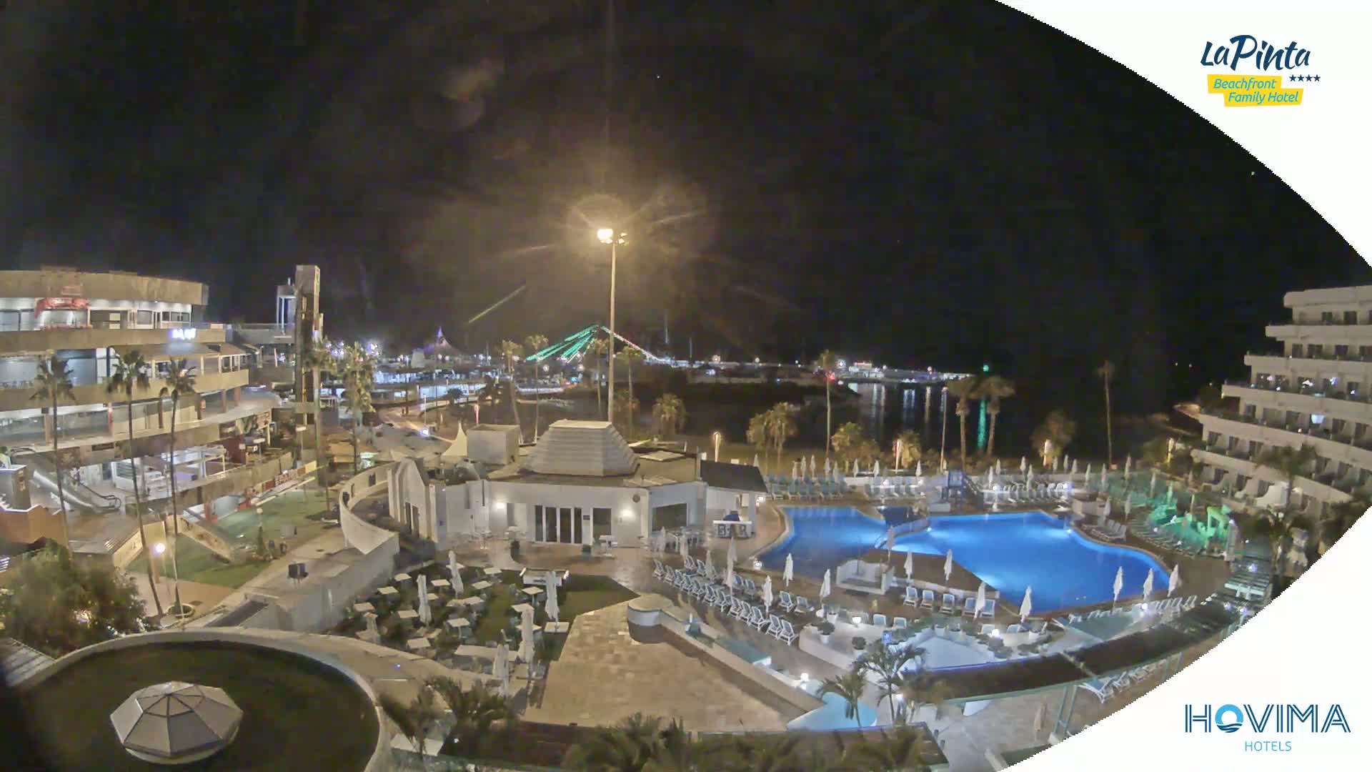 A nighttime aerial view of a resort complex with swimming pools, surrounding buildings, and a waterfront illuminated by lights, under a clear sky.