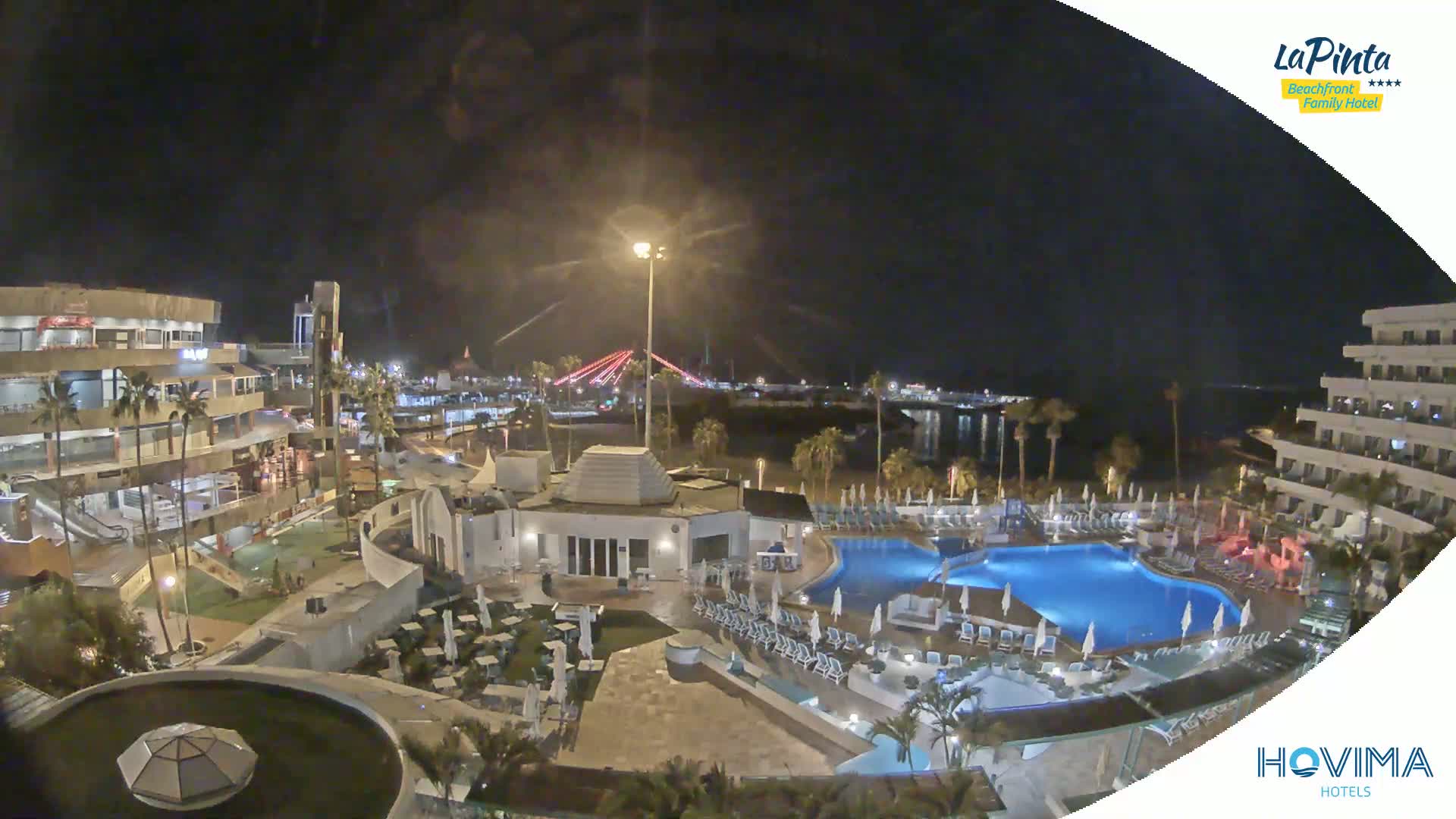 A nighttime view of a resort complex with swimming pools, surrounded by buildings and palm trees, overlooks a calm body of water and a distant bridge under a clear, dark sky.