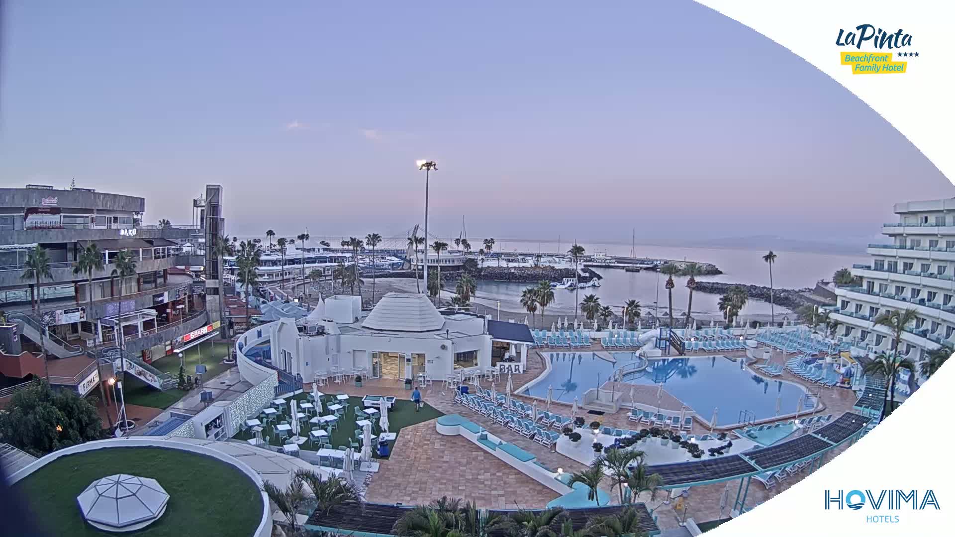 A resort area with a swimming pool and a marina is seen at dawn under a clear, light purple sky.