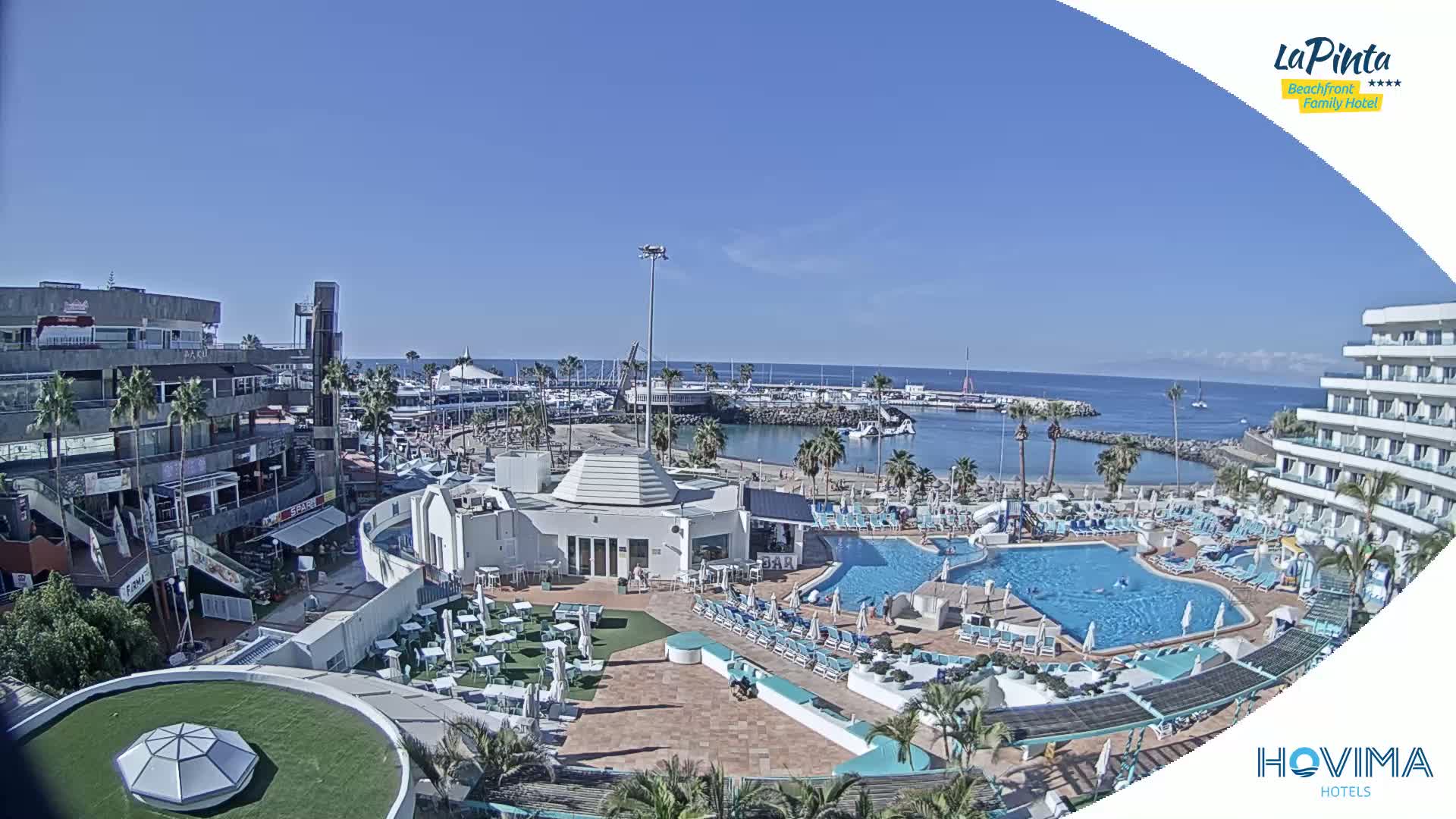 An aerial view shows a resort pool area, adjacent shops and restaurants, a marina filled with boats, and a calm ocean under a clear blue sky.