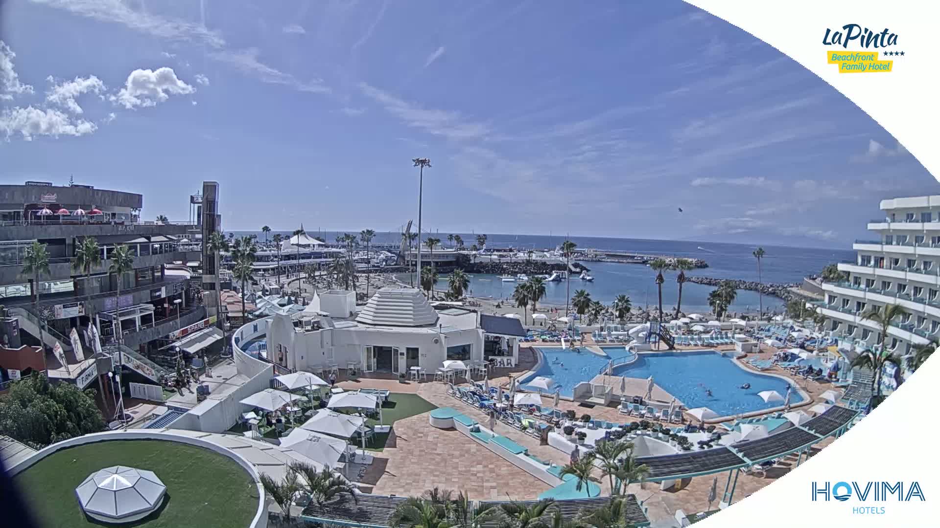 An aerial view shows a resort with a pool and beach area on a sunny day, overlooking a harbor with boats and a calm ocean.