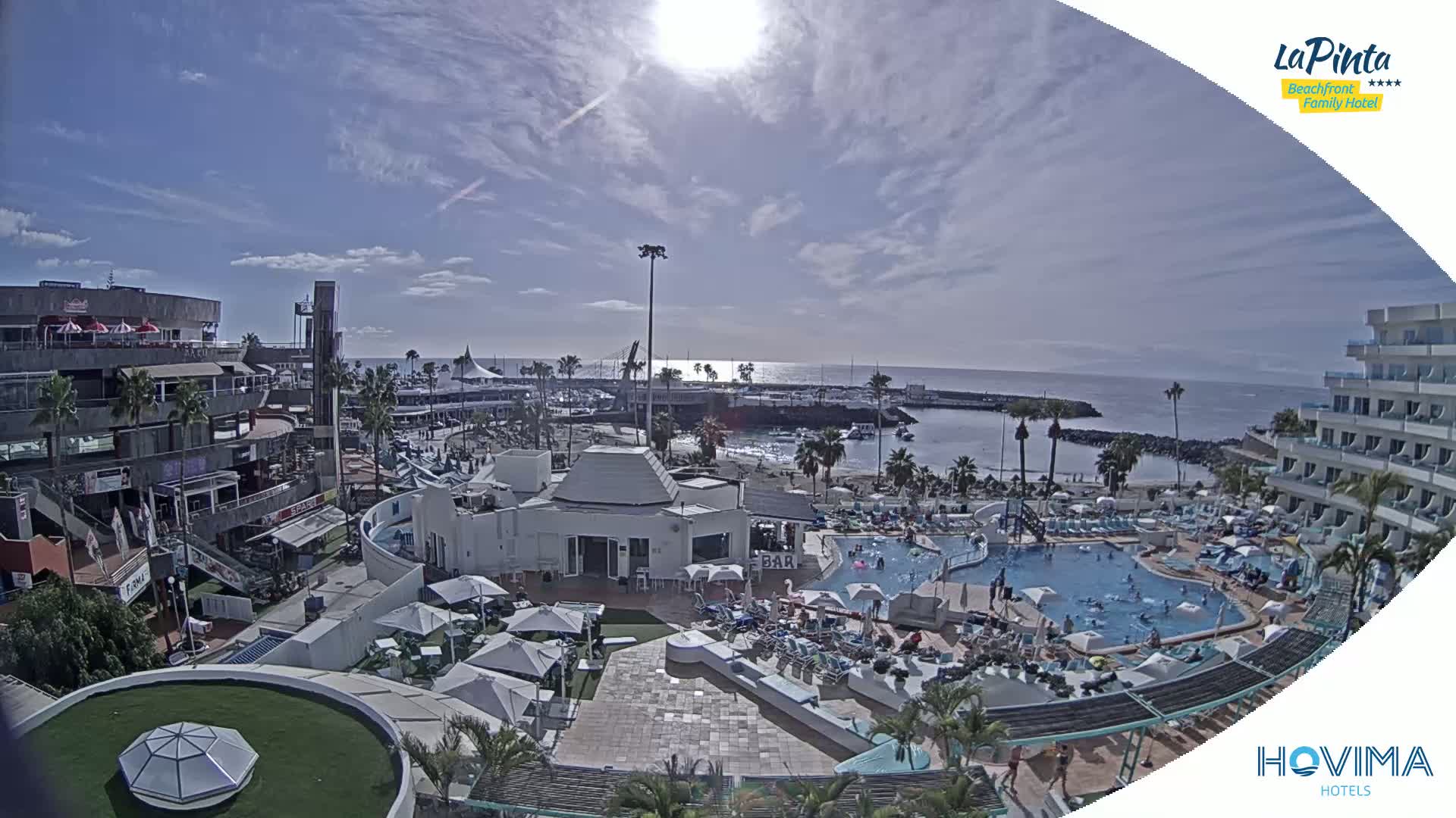 A sunny day overlooks a resort area with pools, buildings, a marina, and ocean views.