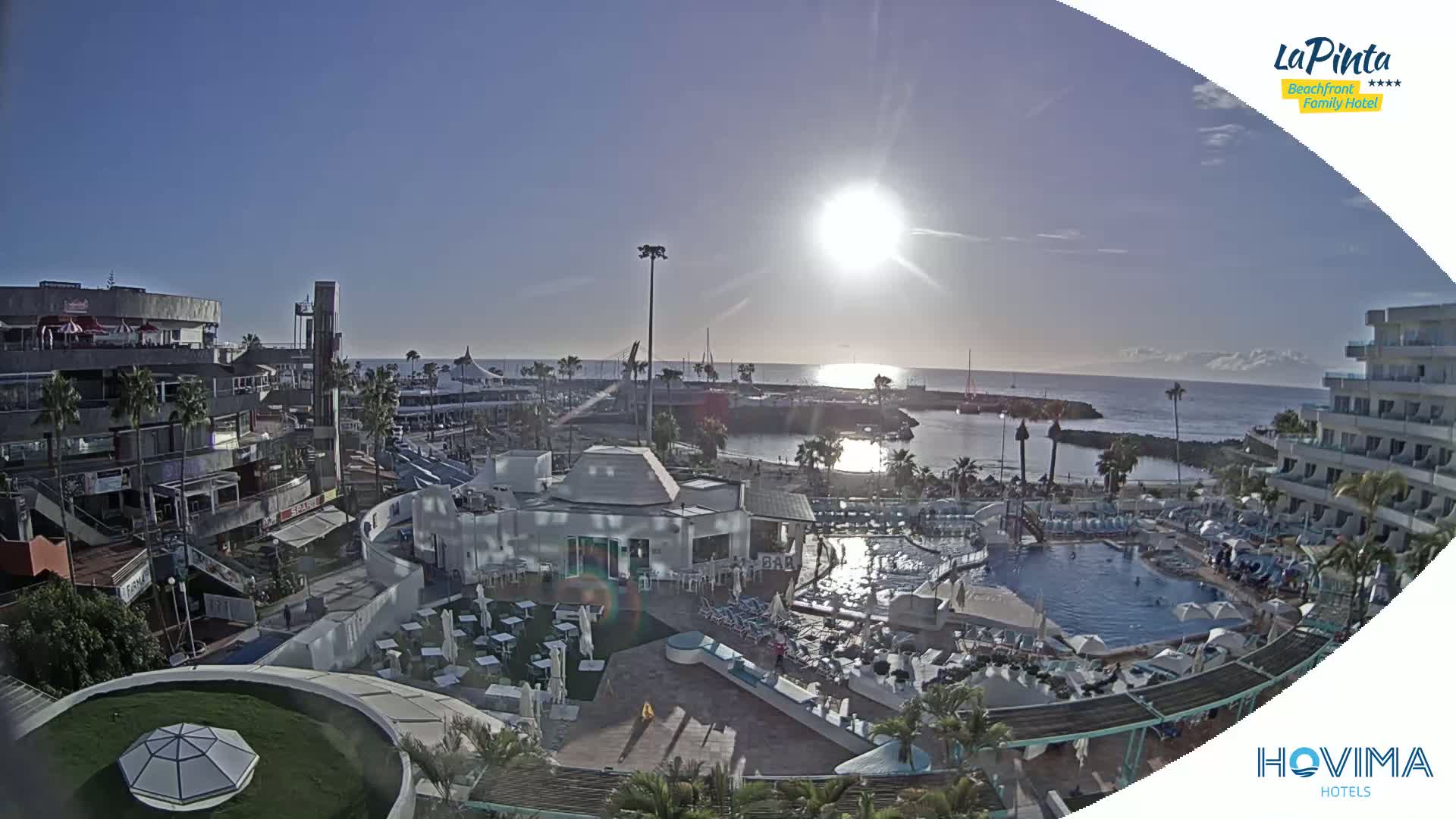A sunny day overlooking a resort pool area, marina, and shops near a calm ocean.