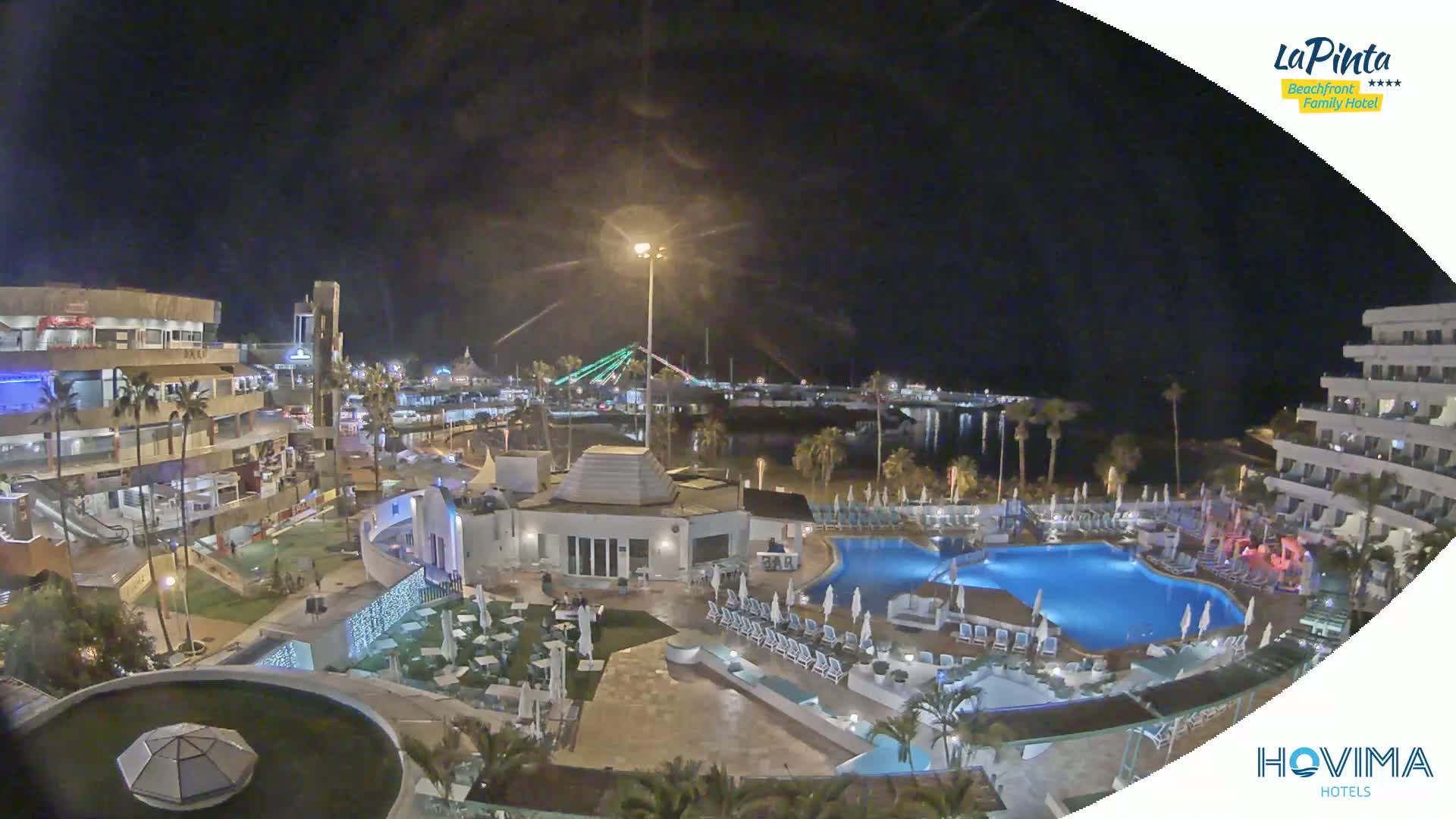 A nighttime aerial view of a resort complex with a pool, situated near a waterfront and illuminated buildings, under a clear night sky.