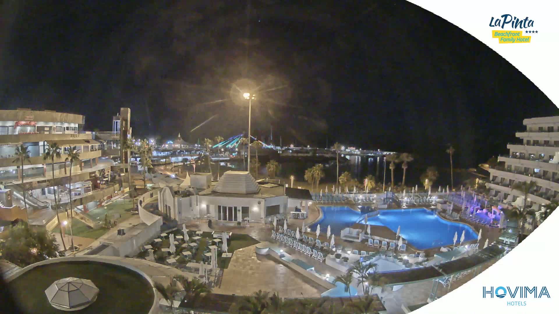 A nighttime aerial view of a resort complex with swimming pools, surrounding buildings, and a waterfront illuminated by lights.