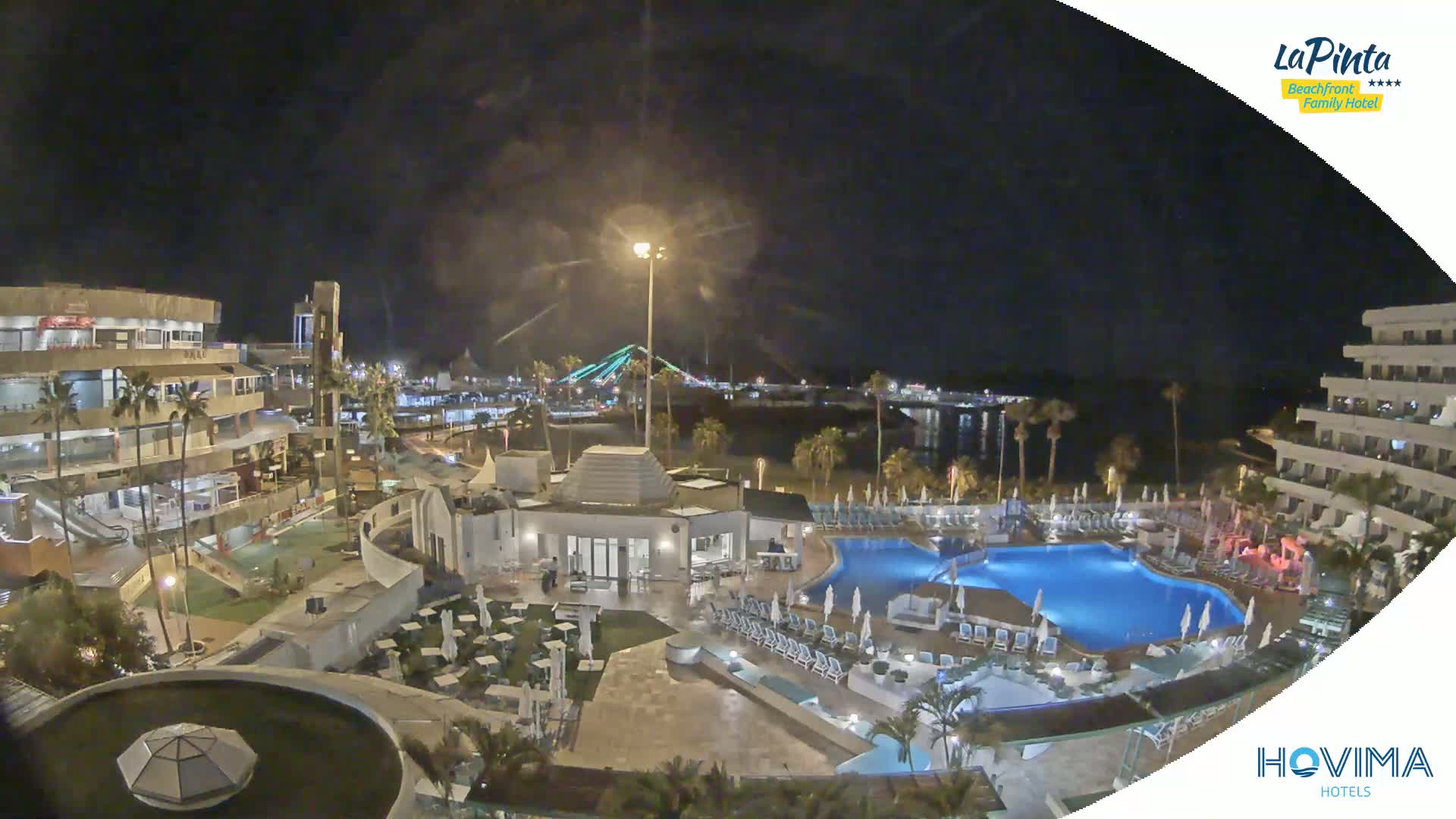 A nighttime aerial view of a resort complex with swimming pools, surrounding buildings, and a bridge in the distance under a clear sky.