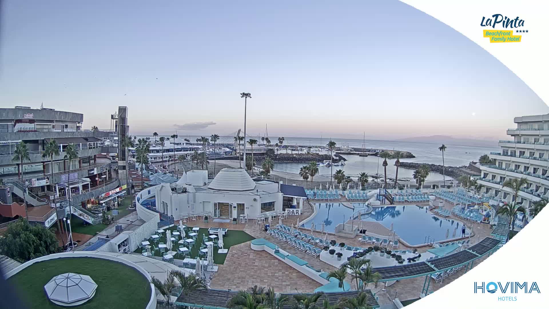 An aerial view of a resort, including a large swimming pool, beach, marina, and buildings, under a clear twilight sky.