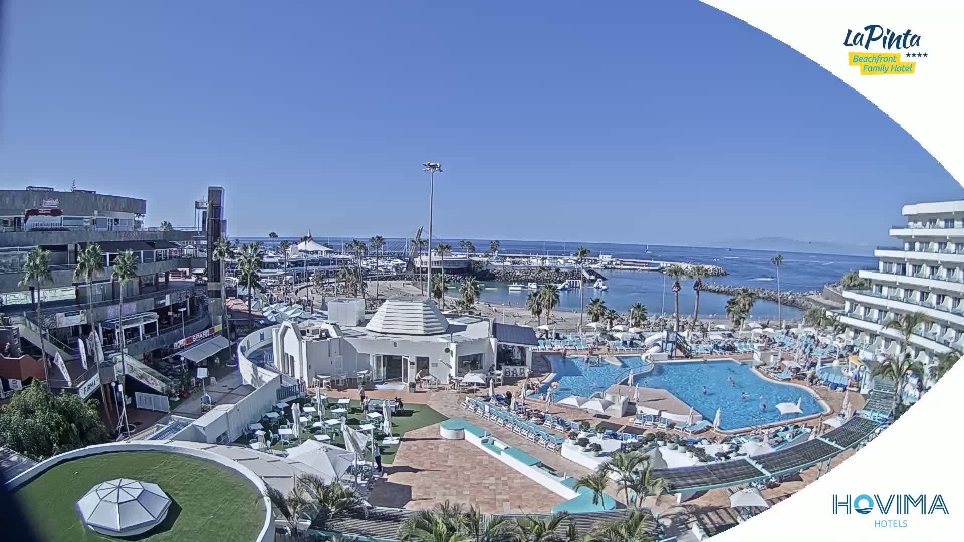 A sunny beach resort with a large pool, adjacent buildings, and a marina in the background.