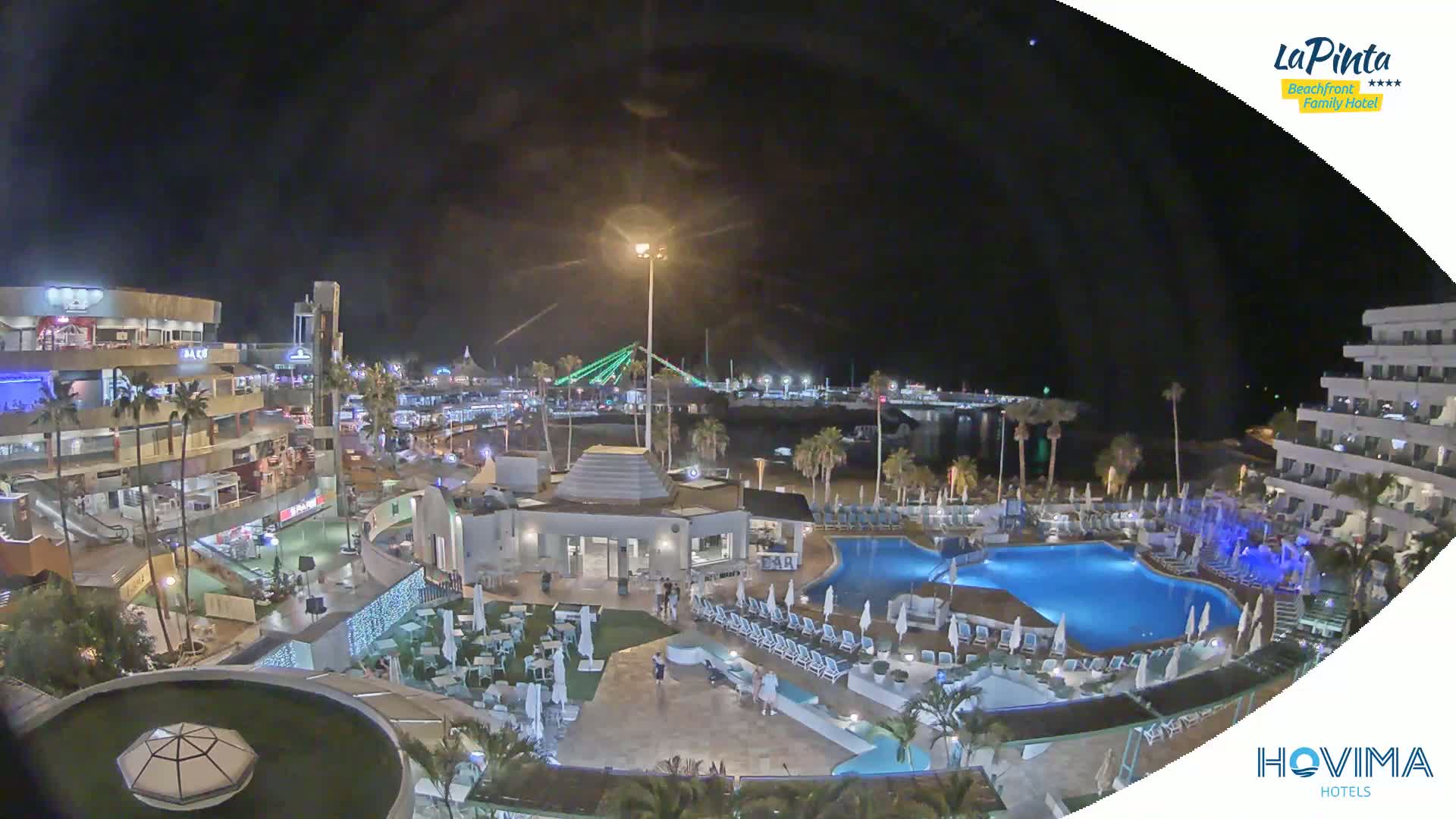 A nighttime aerial view of a resort with a pool and surrounding buildings, overlooking a harbor and a bridge under a clear sky.