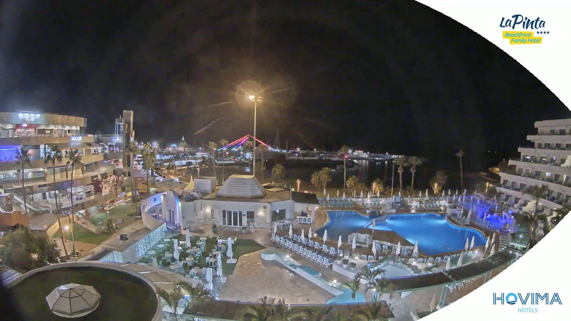 A nighttime aerial view of a resort with a swimming pool complex, adjacent to a waterfront area with shops and a lit bridge.