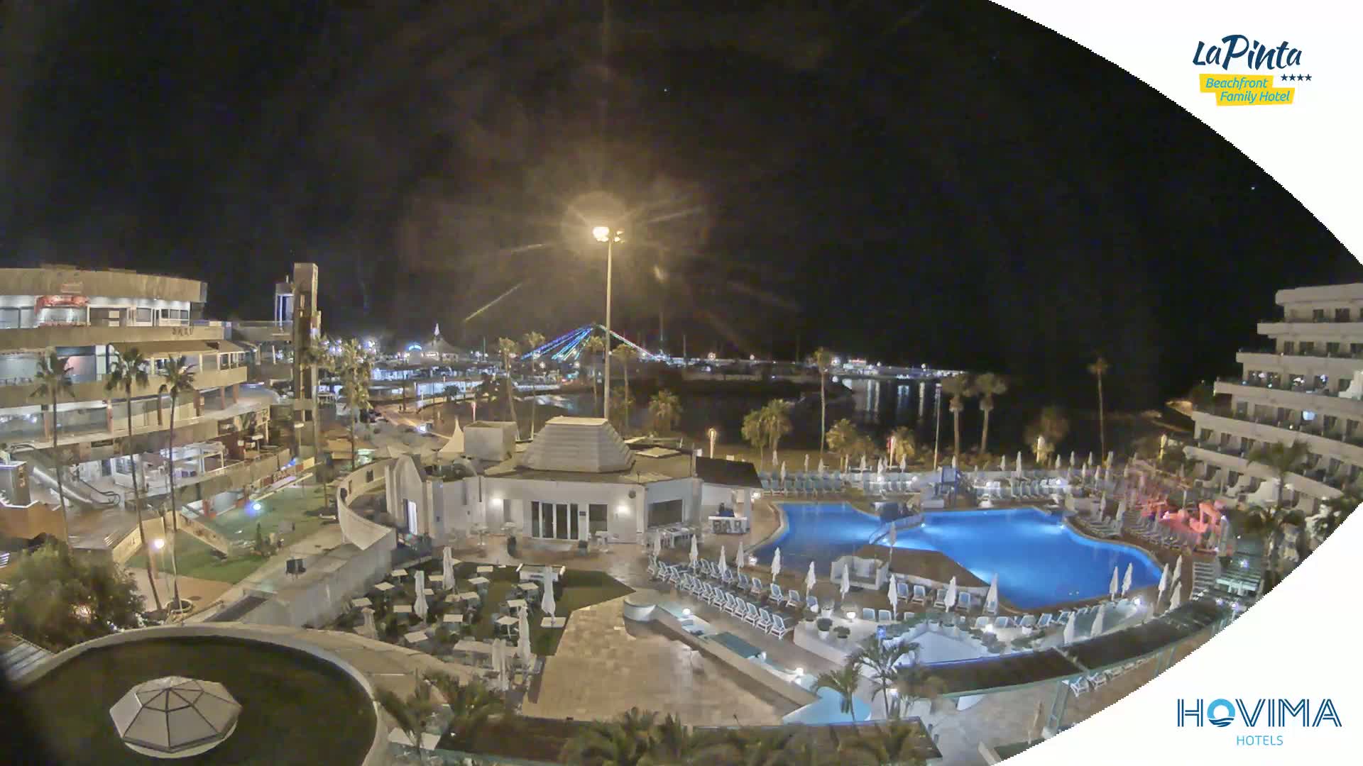 A nighttime aerial view of a resort complex with swimming pools and surrounding buildings, overlooking a calm body of water and a lit-up bridge in the distance.