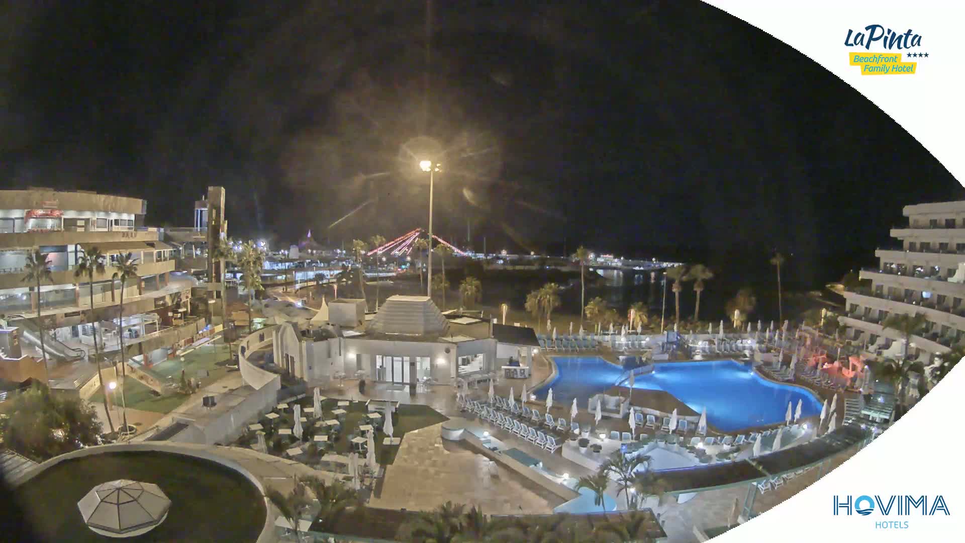 A nighttime aerial view of a resort complex with swimming pools, palm trees, and buildings, overlooking a calm body of water and a distant bridge.
