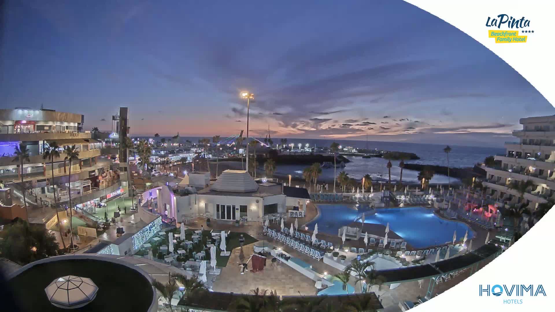 An aerial view of a resort at dusk, showing a swimming pool, surrounding buildings, and a coastline with a calm ocean under a twilight sky.