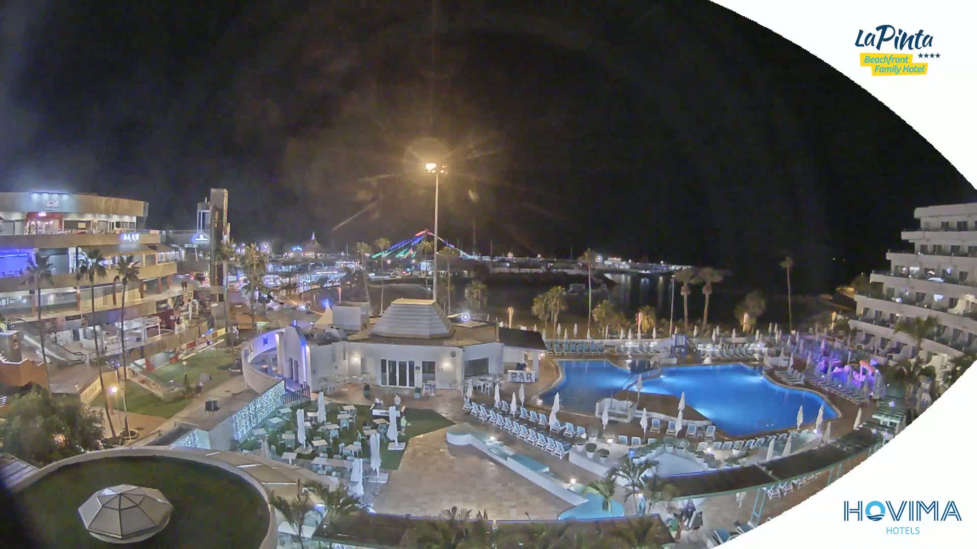 A nighttime aerial view of a resort complex with swimming pools and surrounding buildings, situated near a waterfront with lights visible in the distance under a clear, dark sky.