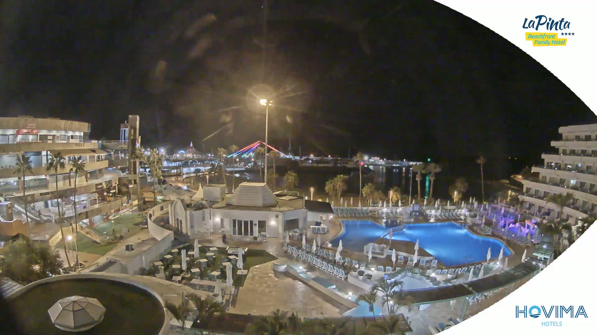 A nighttime view of a resort complex with swimming pools and surrounding buildings, overlooking a calm body of water and a bridge with illuminated lights in the distance.