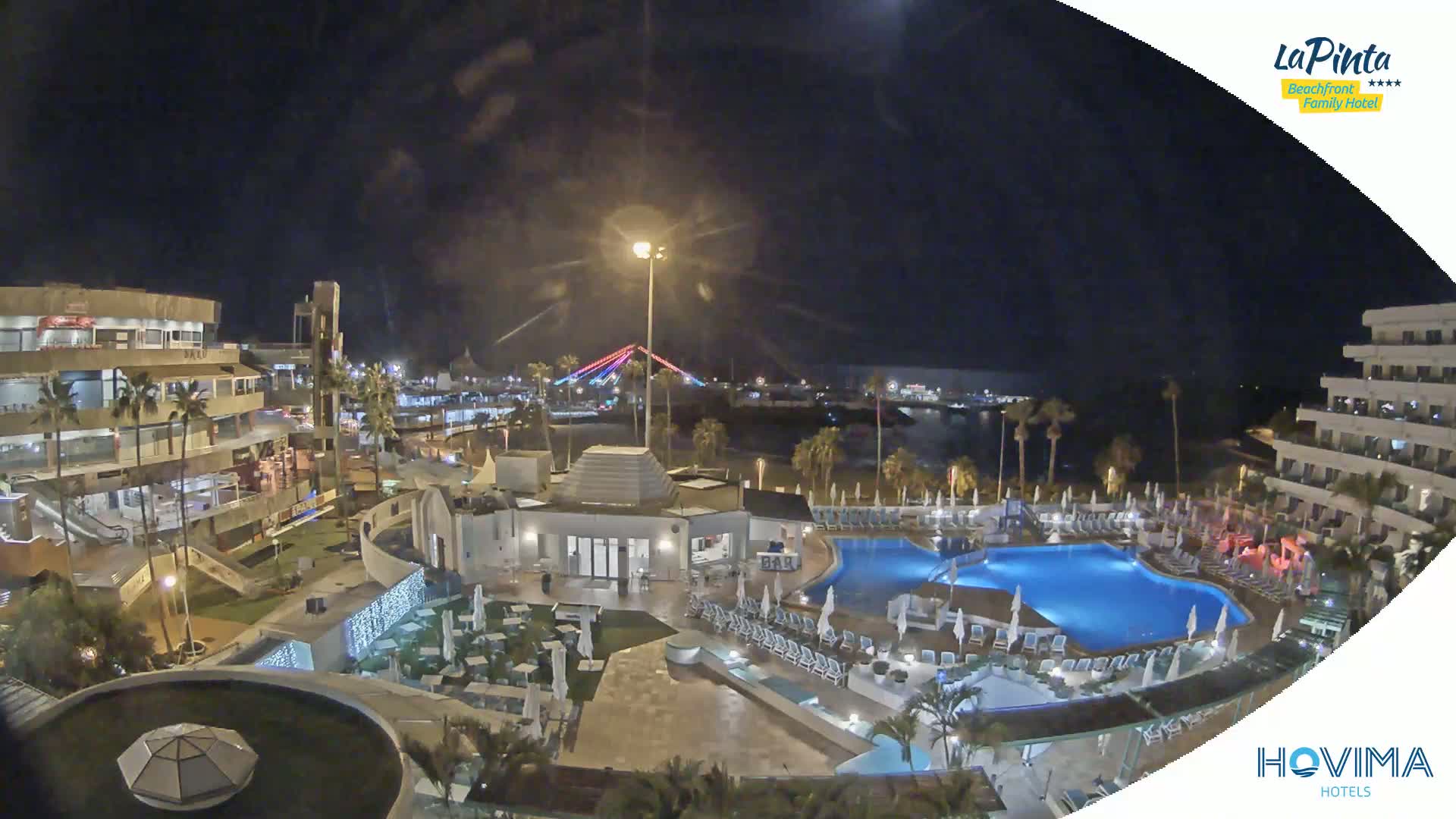 A nighttime aerial view of a resort complex with pools and surrounding buildings, featuring a brightly lit bridge in the distance under a clear, dark sky.