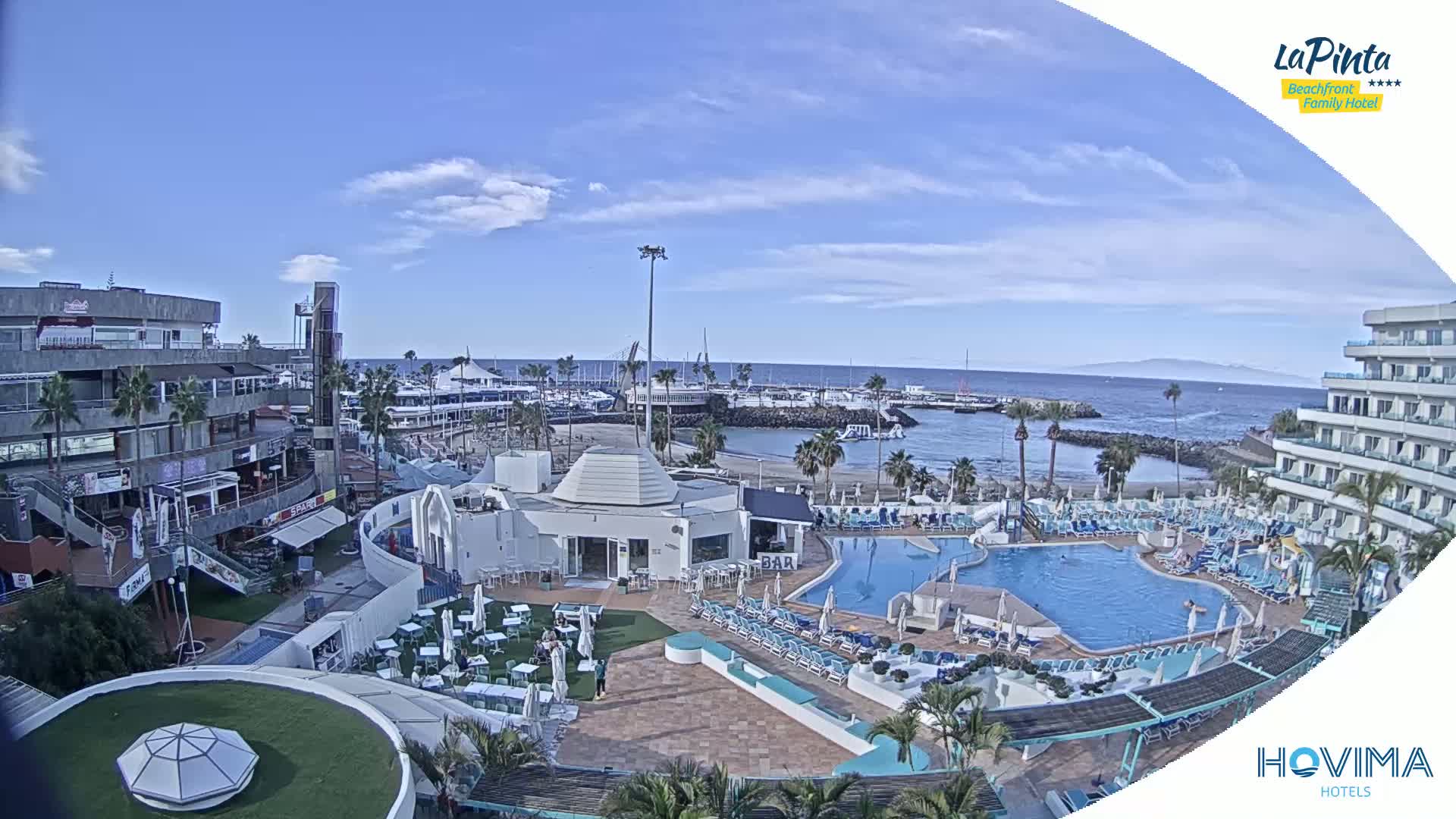 A resort with a swimming pool overlooks a marina and calm ocean under a partly cloudy sky.