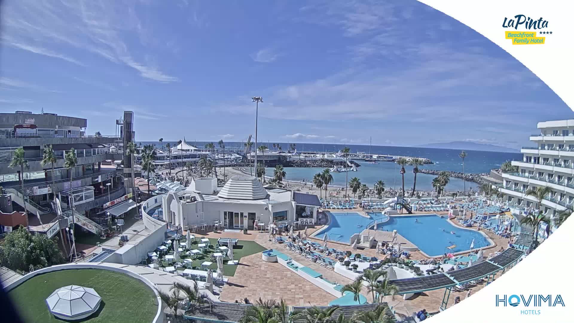 An aerial view shows a resort area with multiple pools, a beach, a marina, and buildings under a mostly sunny sky.