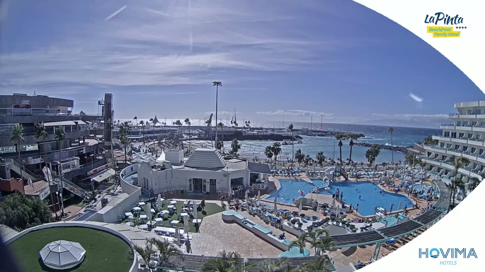 A sunny, seaside resort with multiple pools, a beach, and a harbor is seen from an elevated viewpoint.
