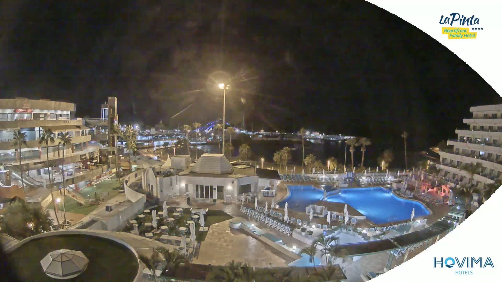 A nighttime aerial view of a resort complex with swimming pools, palm trees, and buildings near a body of water under a clear, dark sky.
