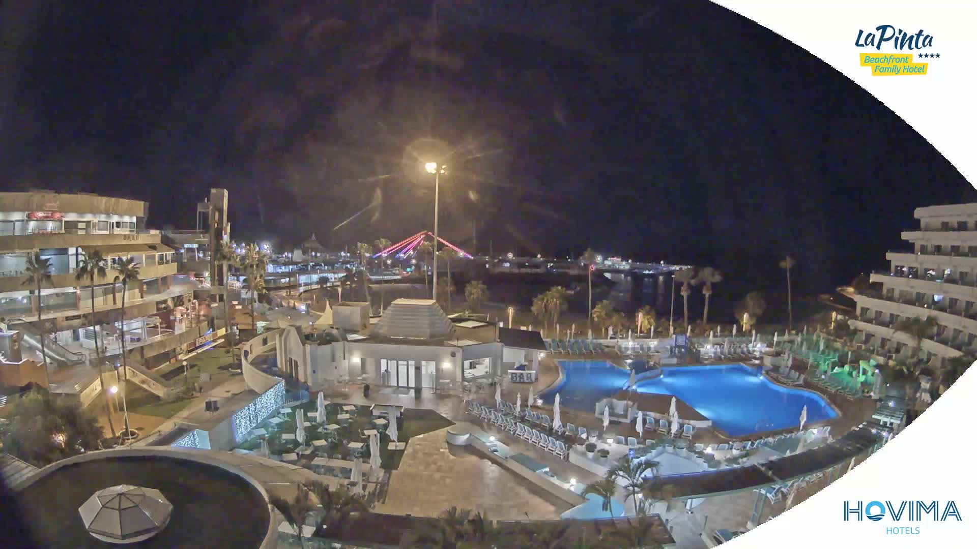 A nighttime aerial view of a resort complex with swimming pools and surrounding buildings, under a clear, dark sky.