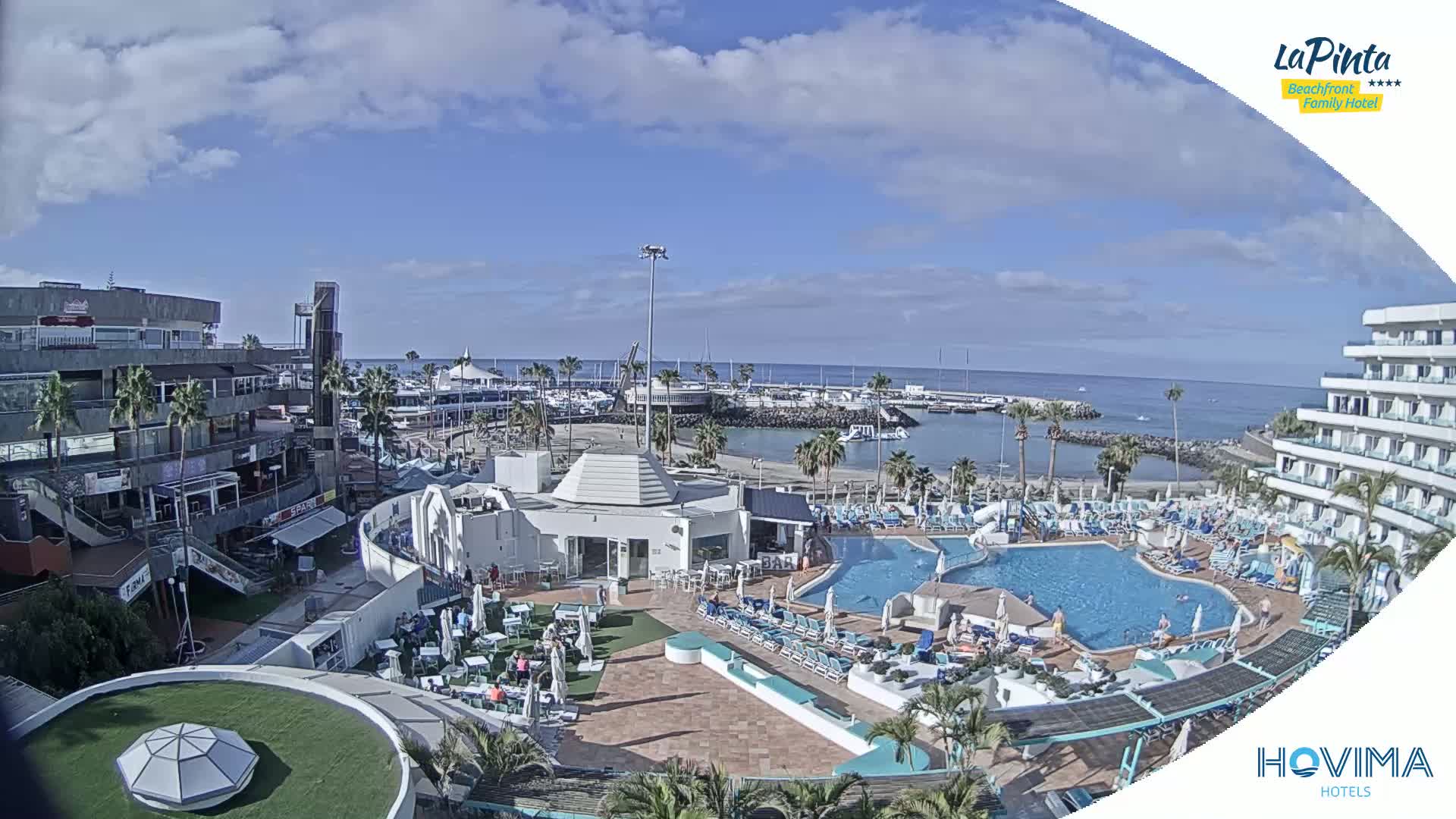 A resort with a swimming pool and surrounding buildings overlooks a marina and ocean under a partly cloudy sky.