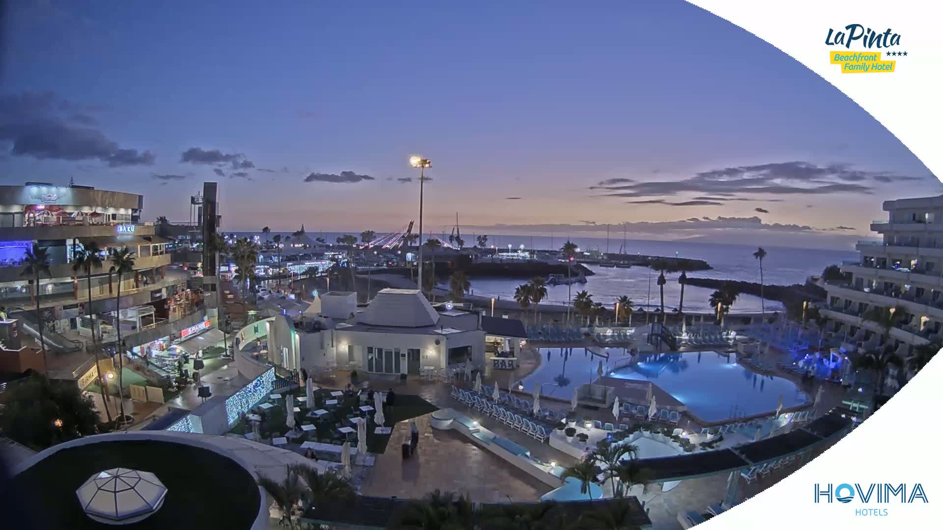 An illuminated resort complex overlooks a calm ocean at twilight, with a marina and town visible in the background.