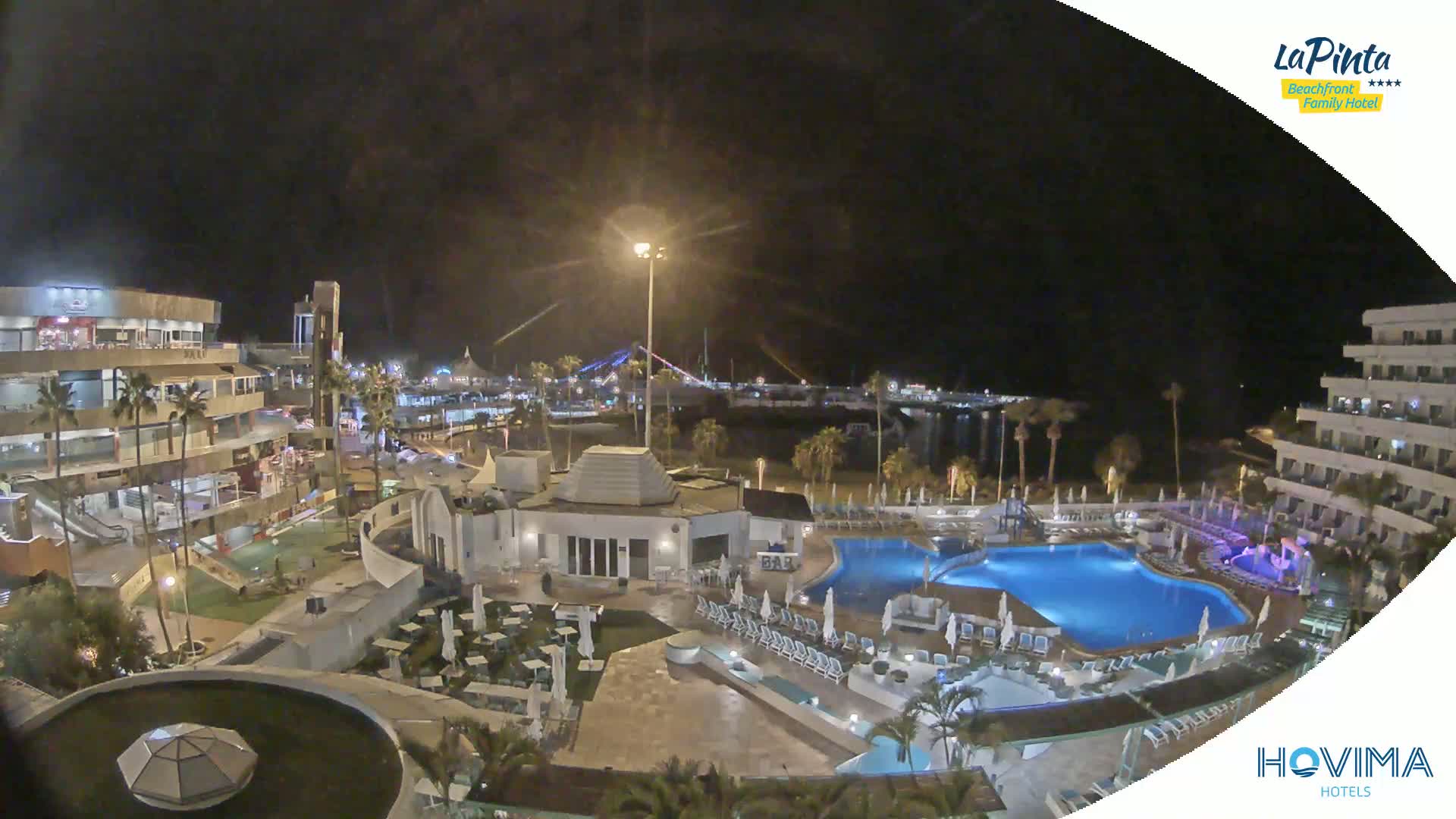 A nighttime aerial view of a resort complex with pools and surrounding buildings, illuminated under a clear, dark sky.