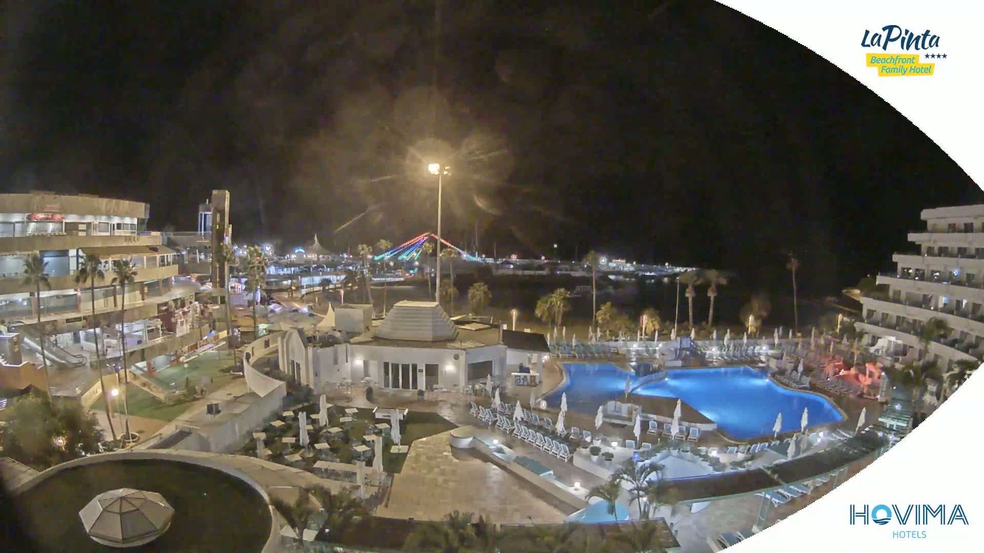 A nighttime aerial view of a resort complex with a swimming pool, surrounding buildings, and palm trees, overlooking a brightly lit beach area and a bridge in the distance.