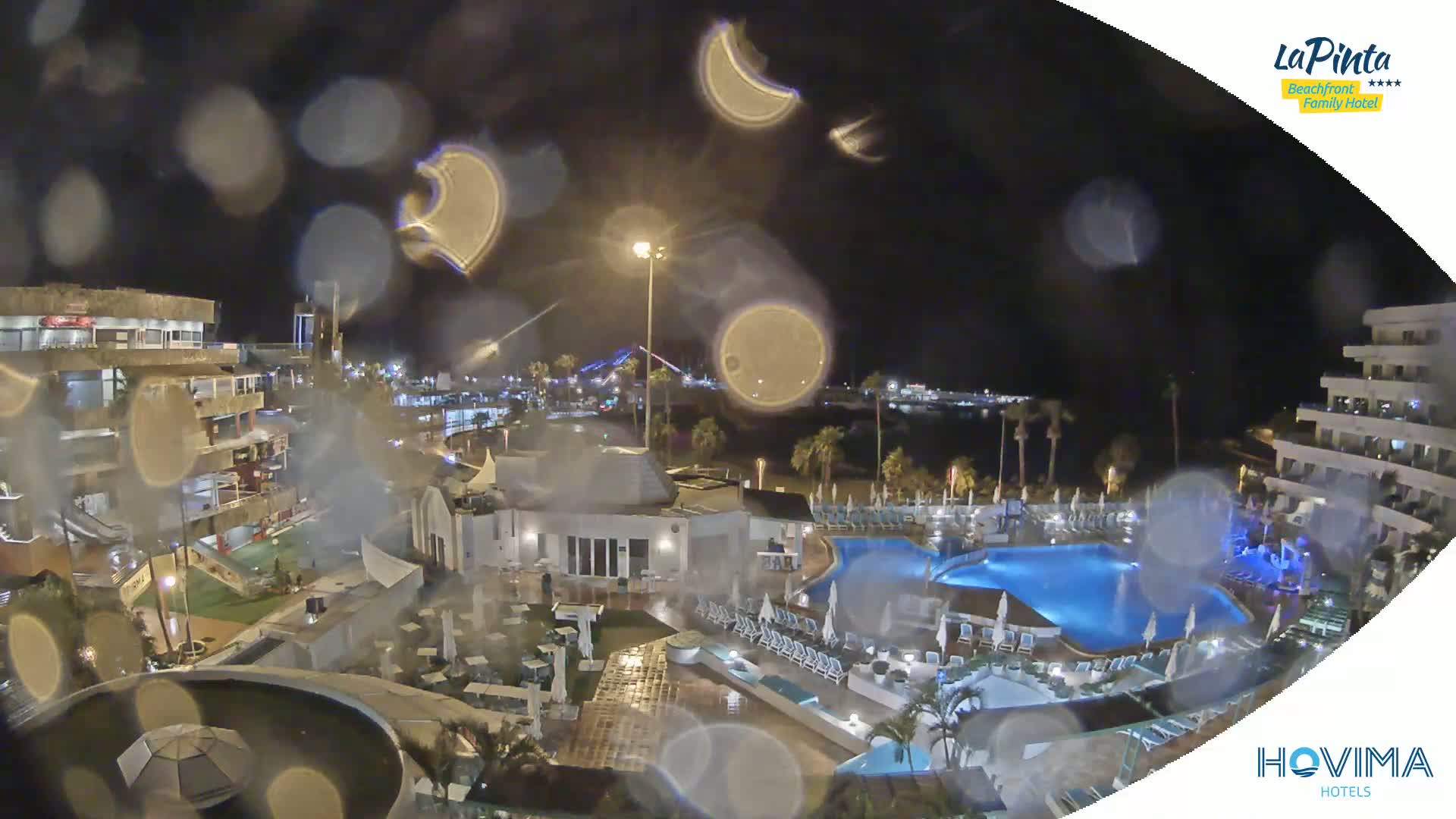 A nighttime view of a resort complex with swimming pools, seen through rain-covered glass, showing palm trees and a body of water in the distance.