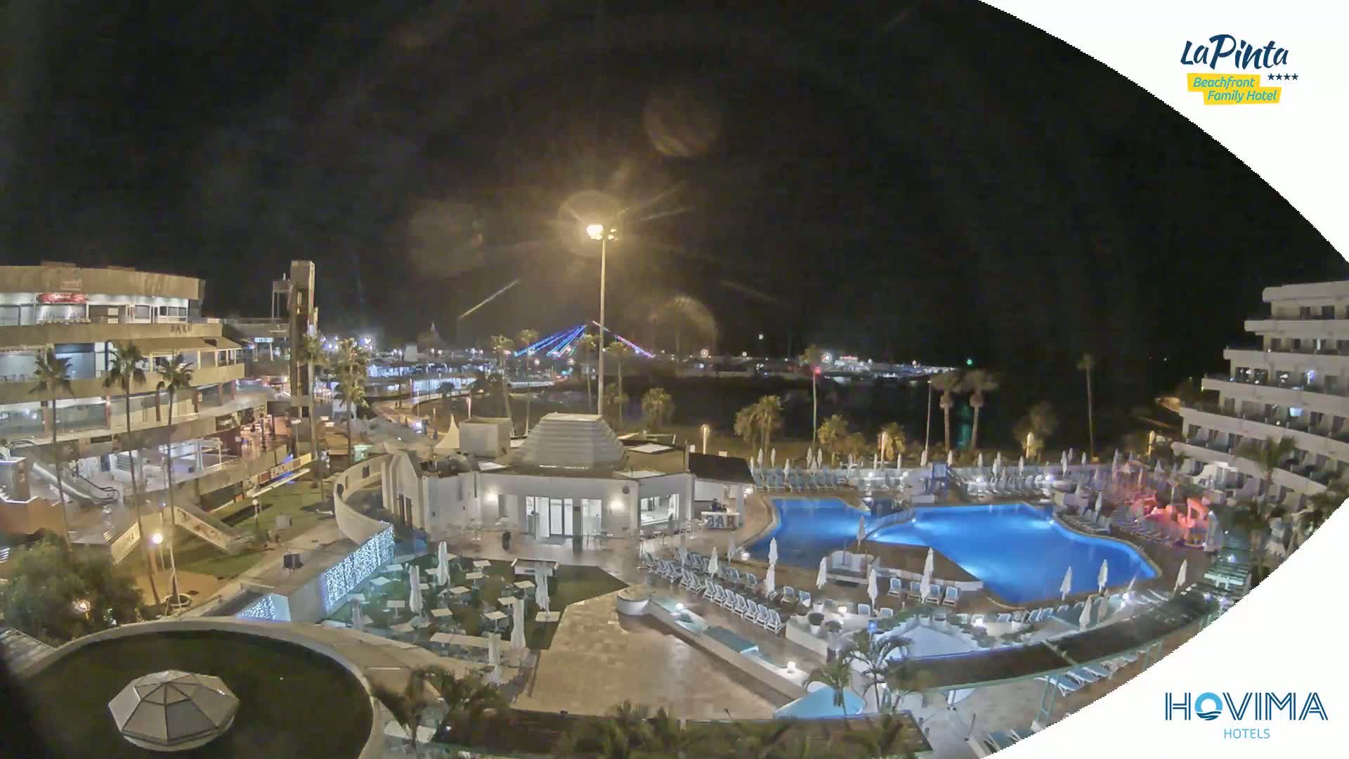 A nighttime aerial view of a resort complex with a swimming pool, surrounding buildings, and a brightly lit bridge in the distance.
