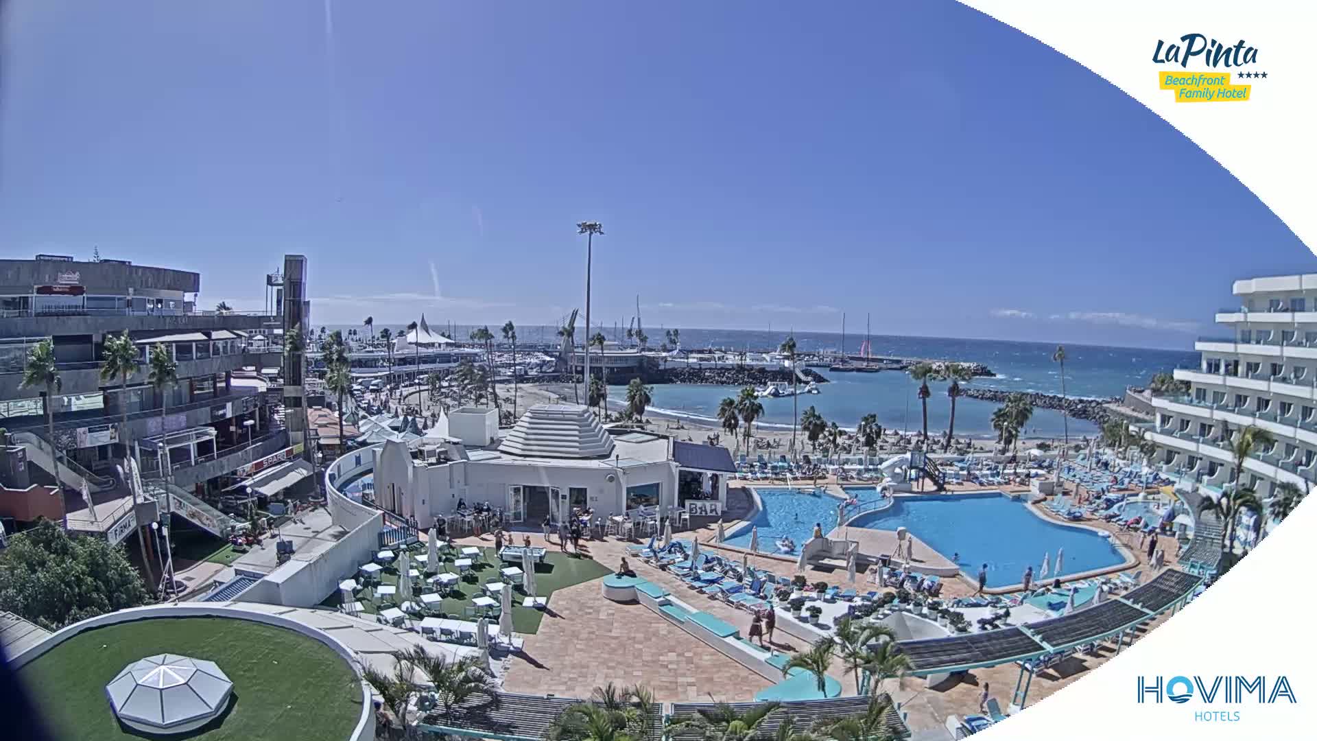 A resort area with pools and a beach, situated next to a marina and town, under sunny skies.