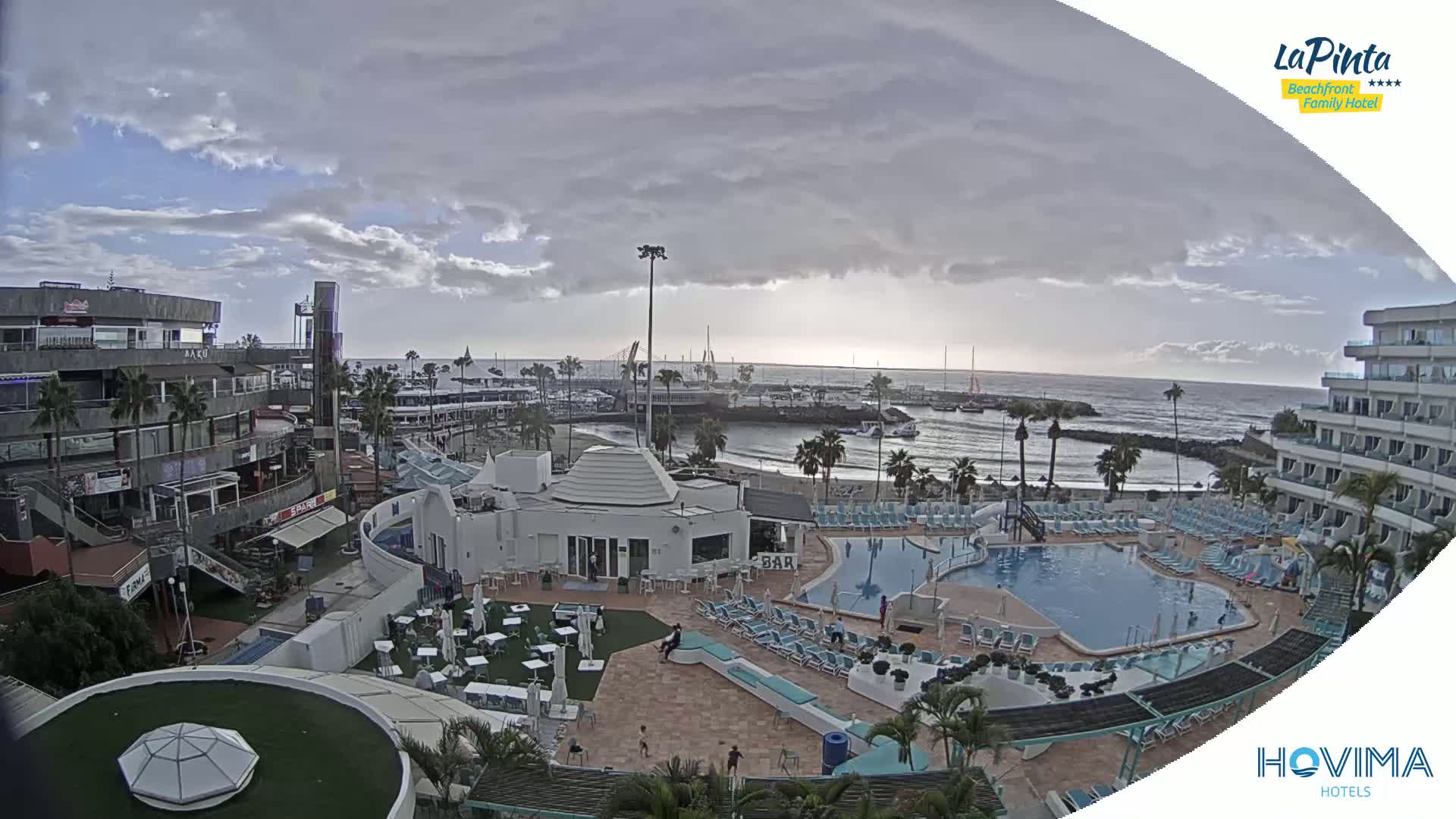 An overcast day shows a resort with multiple pools, a beach, a harbor filled with boats, and buildings with shops and restaurants.