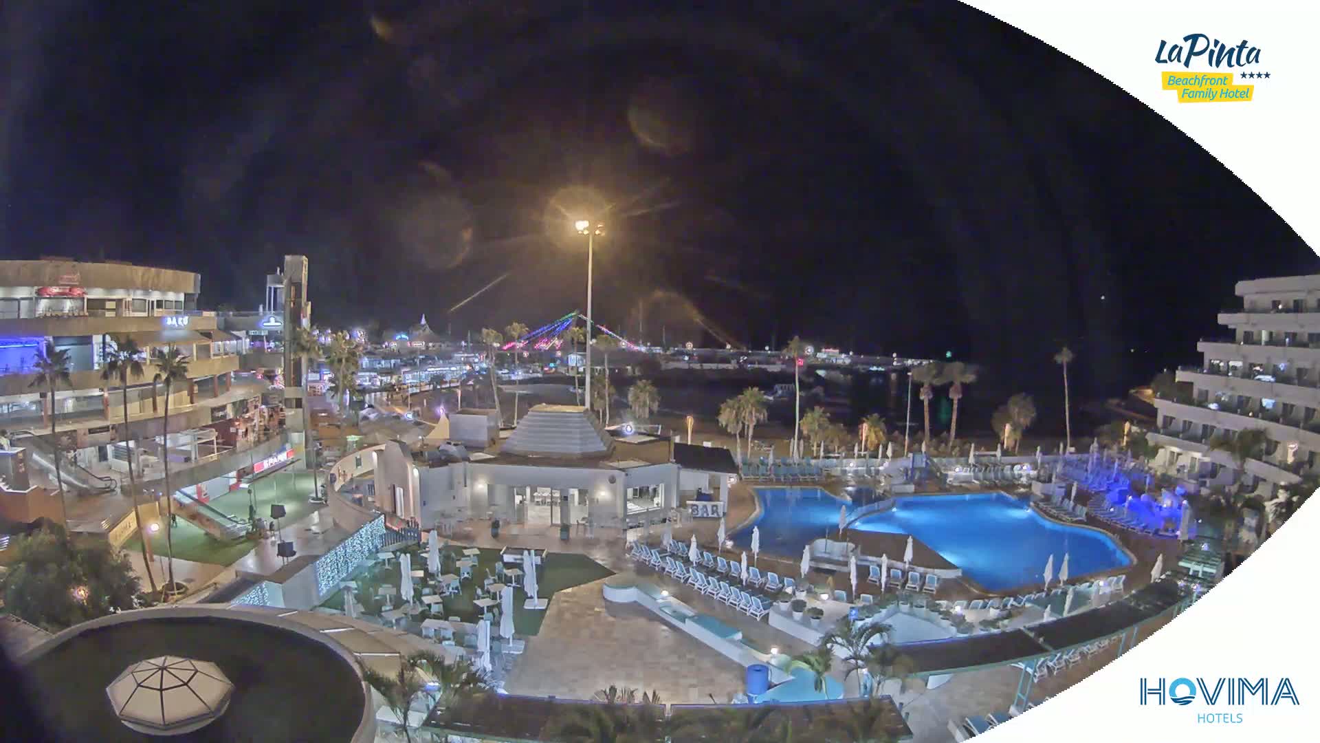 A nighttime aerial view of a resort complex with swimming pools, palm trees, and a beachfront area, under a clear, dark sky.