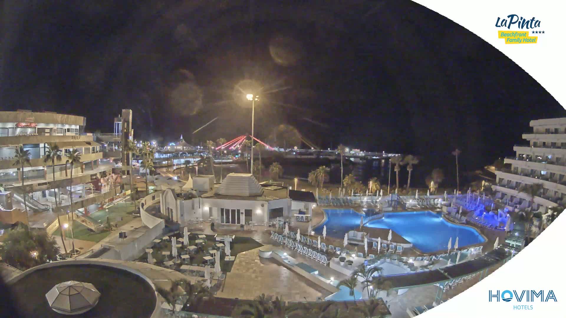 A nighttime aerial view of a resort complex with swimming pools, surrounded by buildings and palm trees, overlooks a beach and a bridge illuminated with red lights.