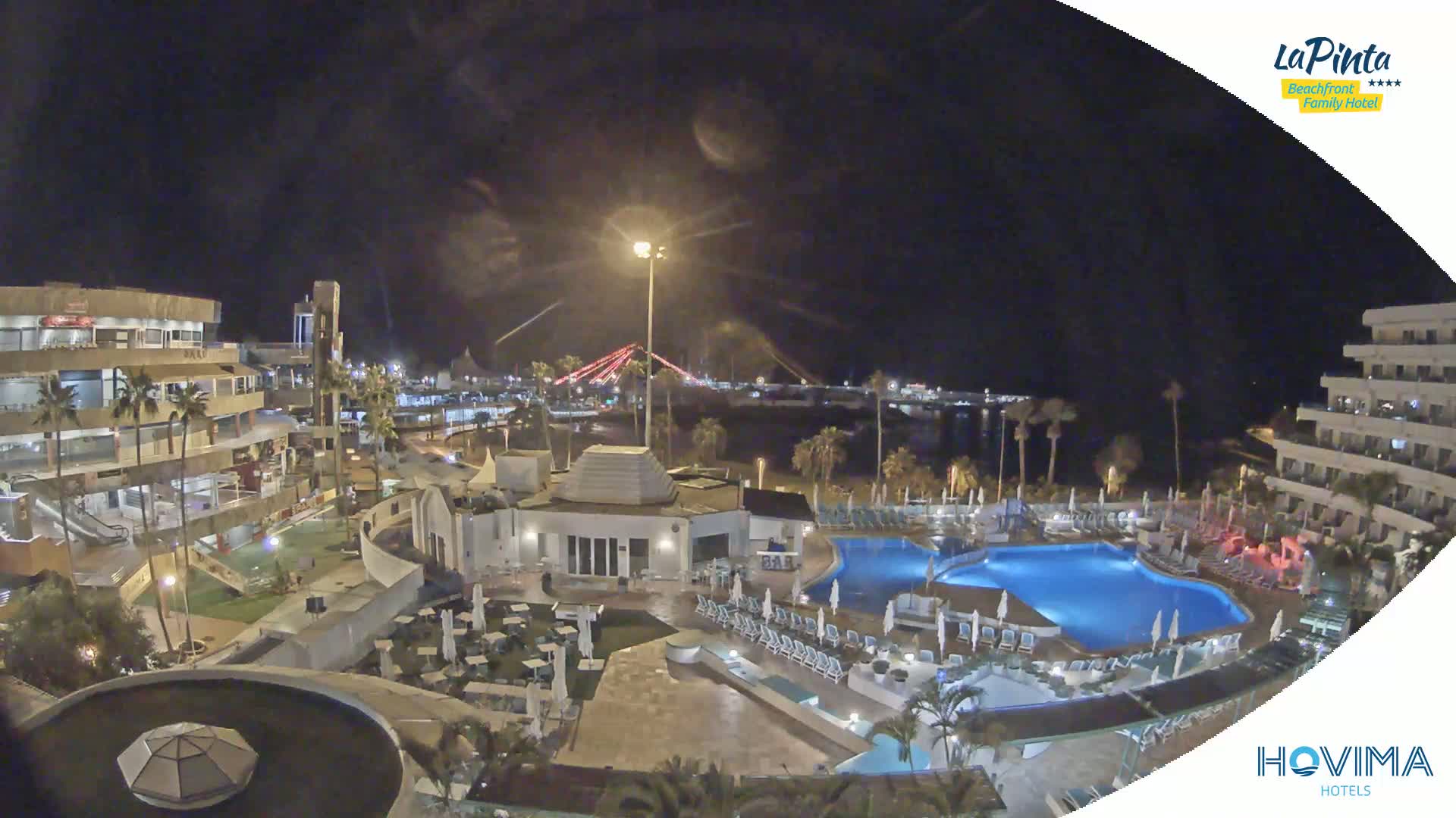 A nighttime view of a resort complex with swimming pools, surrounded by buildings and palm trees, overlooking a calm body of water and a bridge in the distance.
