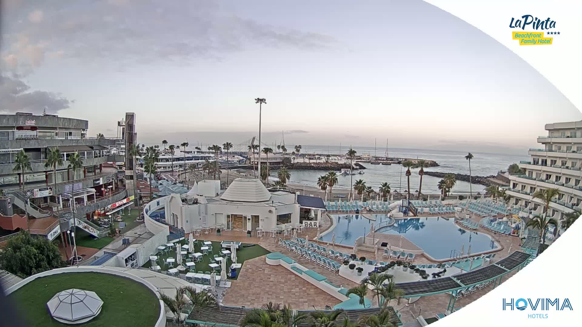 An aerial view of a resort complex with a swimming pool and beach access, overlooking a calm ocean under an overcast sky.