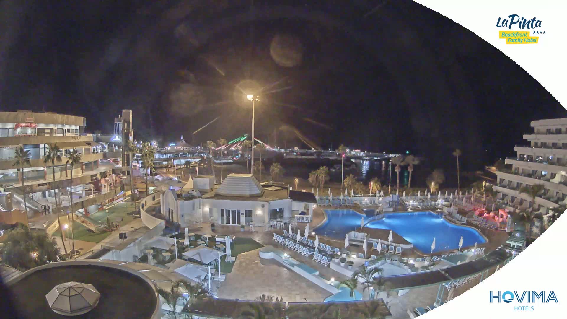 A nighttime aerial view of a resort complex with swimming pools, surrounding buildings, and a beach in the background under a clear, dark sky.