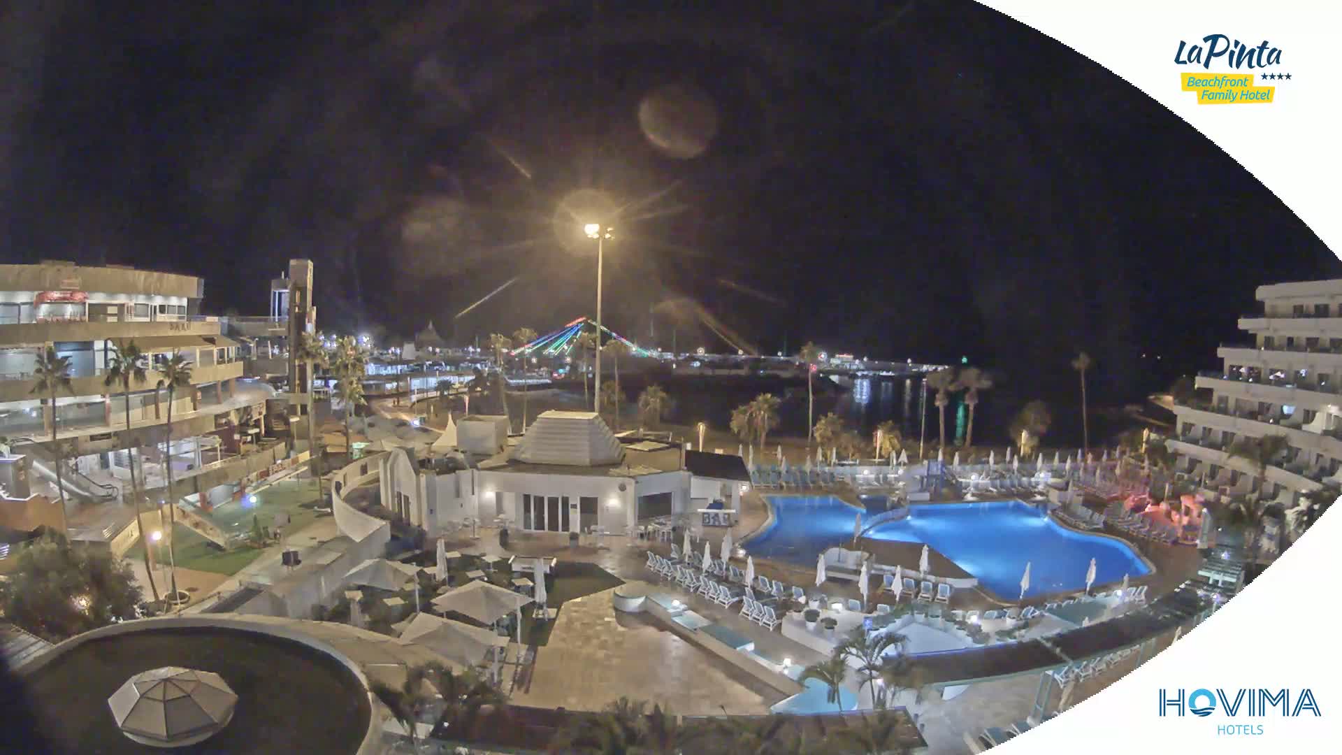 A nighttime aerial view of a resort complex with swimming pools, surrounding buildings, and a brightly lit waterfront in the distance under a clear sky.