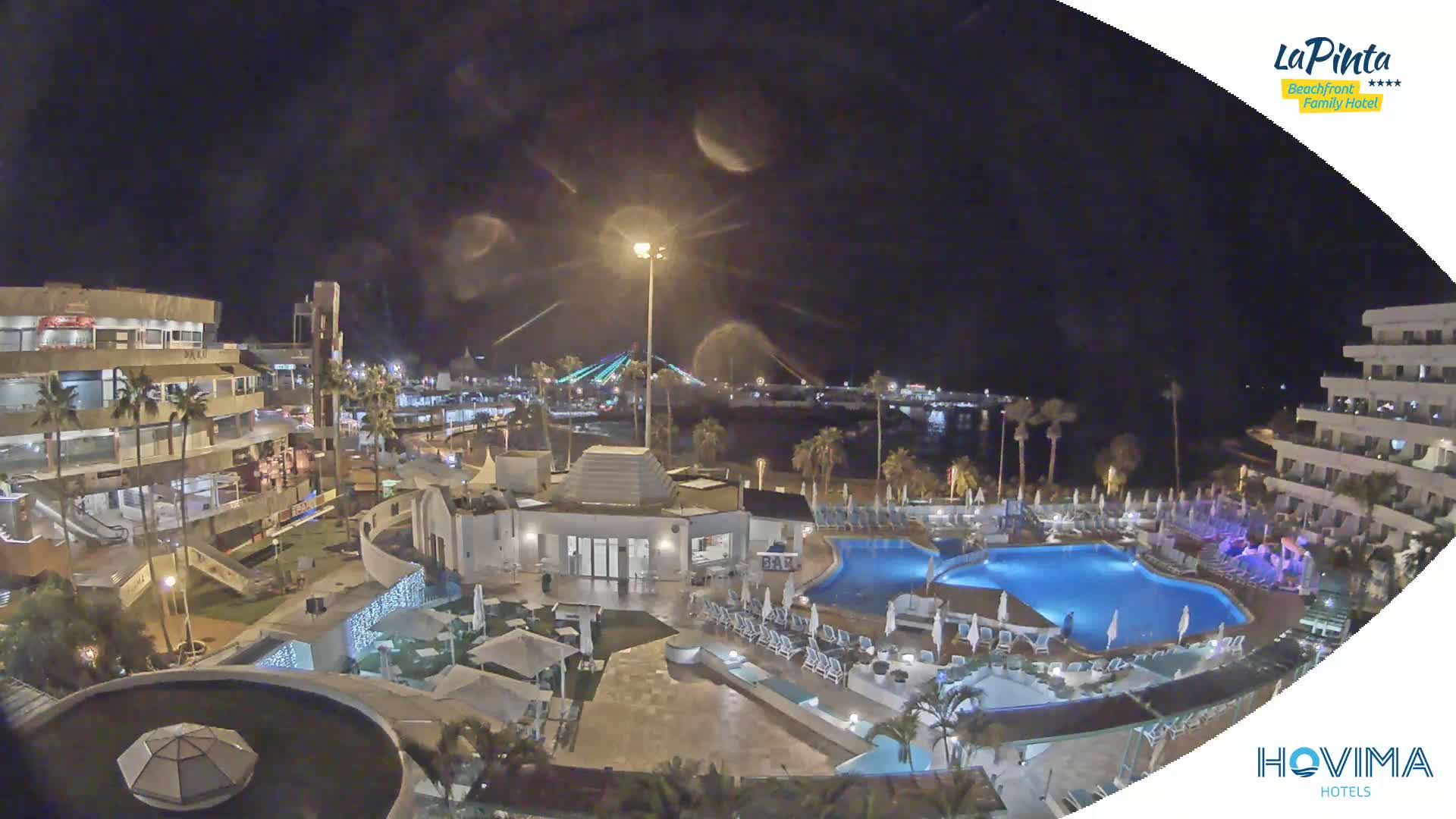 A nighttime aerial view of a resort complex with swimming pools, palm trees, and a nearby waterfront illuminated by lights.
