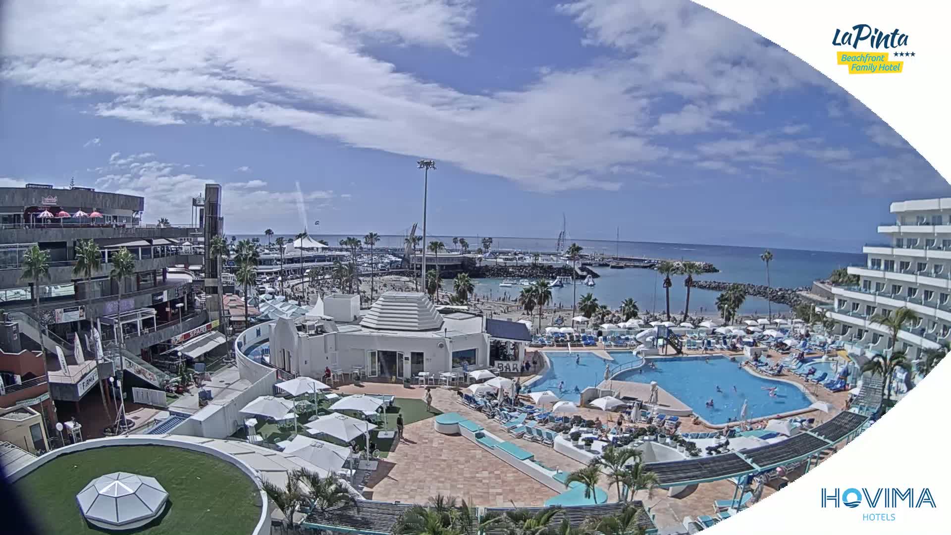 A resort pool area with a beach and marina in the background under partly sunny skies.