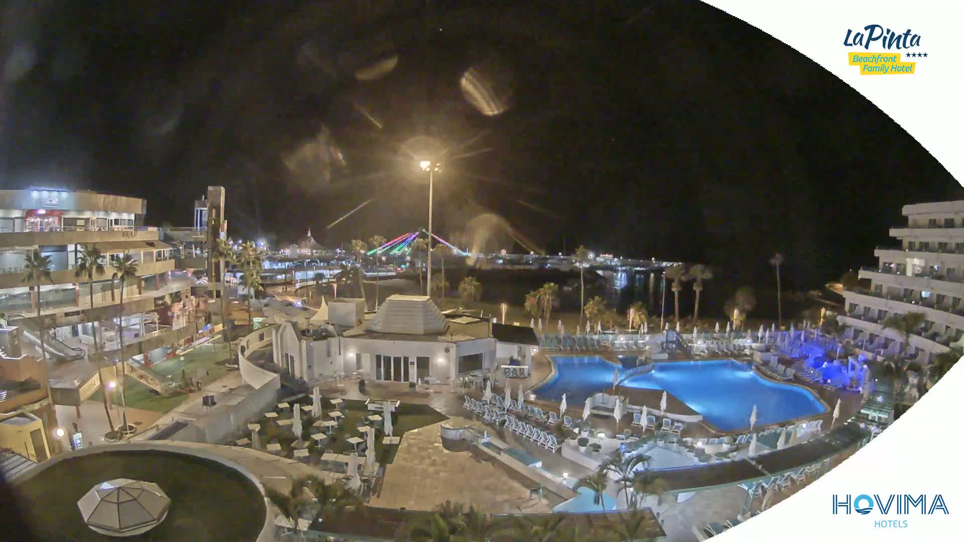 A nighttime aerial view of a resort with swimming pools, palm trees, and a brightly lit boardwalk in the distance under a cloudy sky.