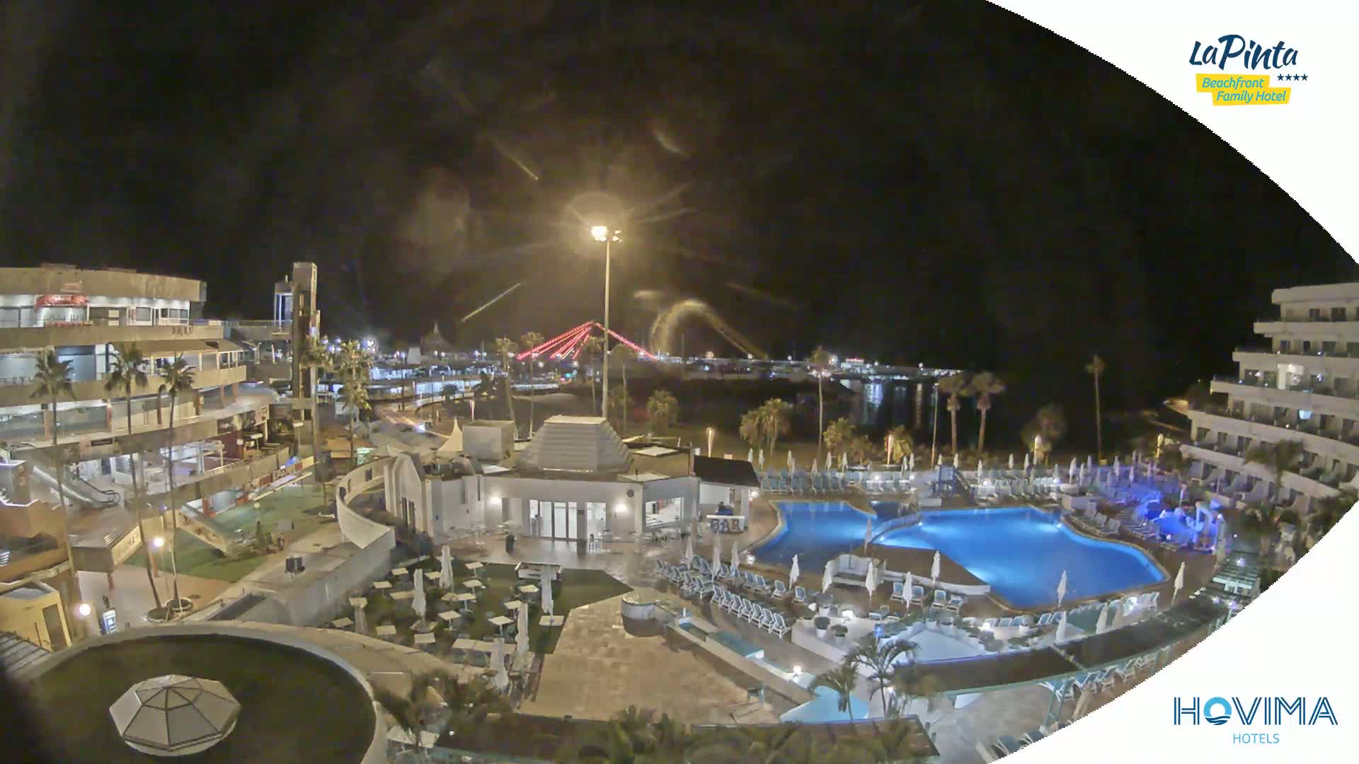 A nighttime aerial view of a resort complex with swimming pools, surrounding buildings, and a beach, illuminated under a partly cloudy sky.