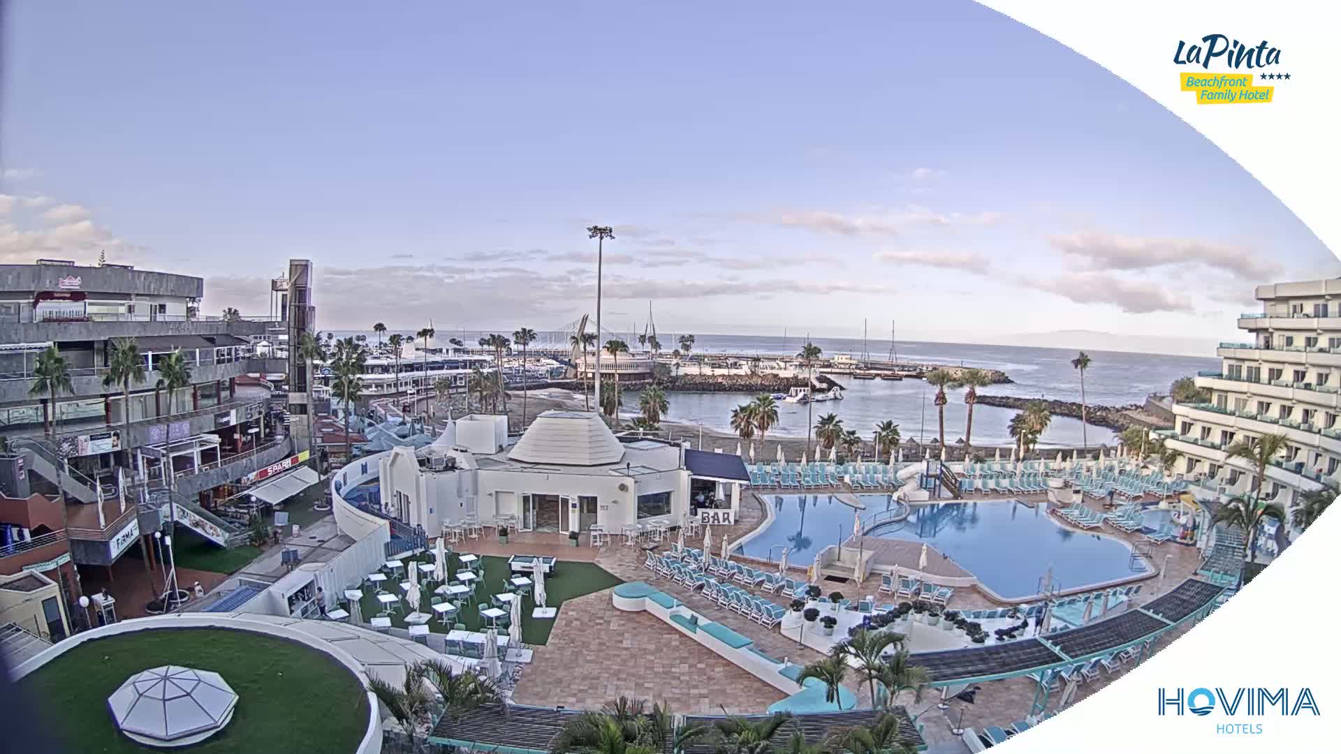 An aerial view of a resort area with a swimming pool, shops, and a marina under a mostly clear sky.