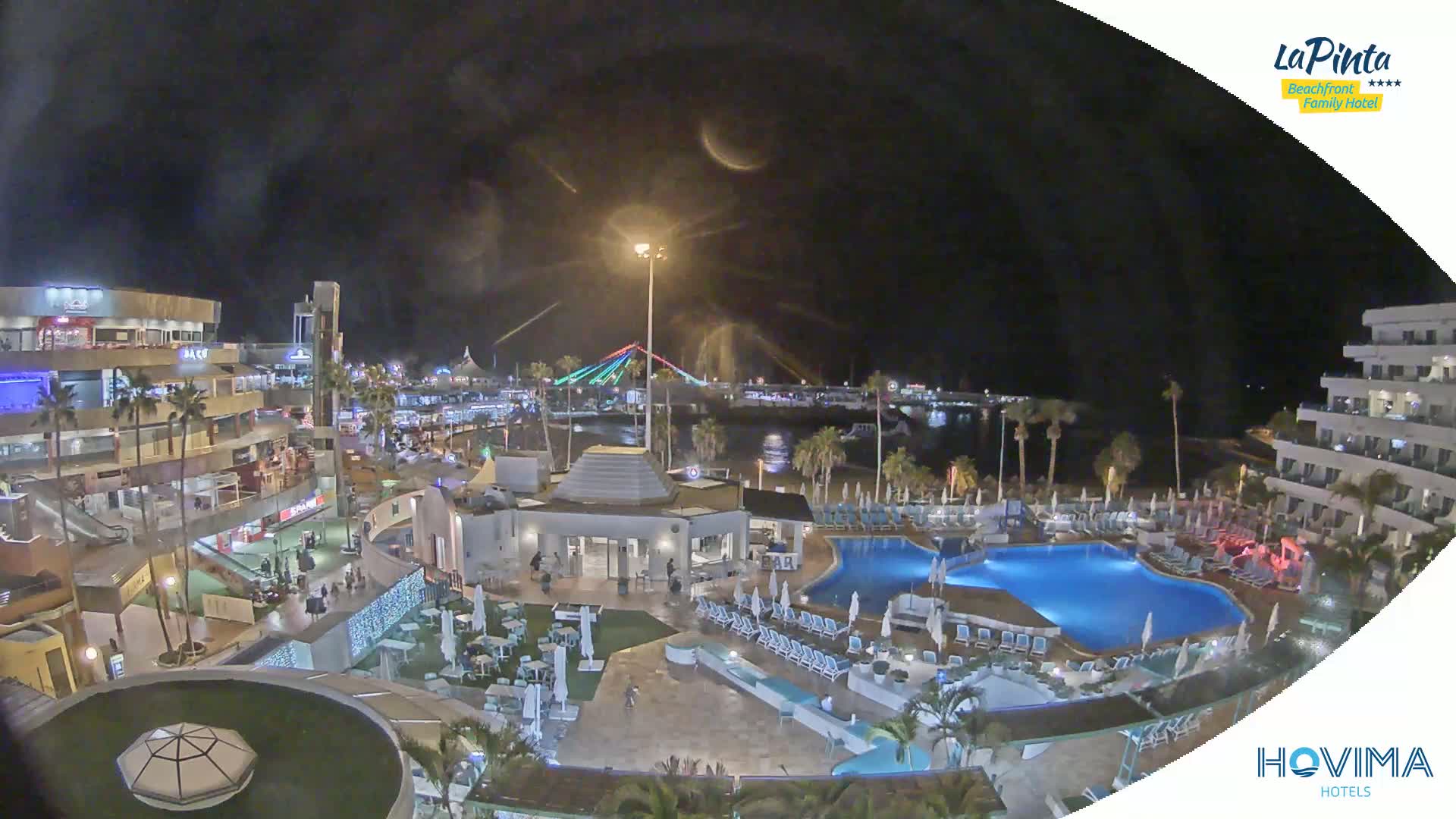 A nighttime aerial view of a resort complex with a swimming pool, surrounded by buildings and palm trees, overlooking a harbor with a lit bridge in the background under a dark, cloudy sky.